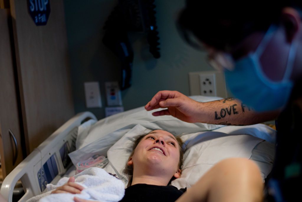 Nurse touches a patient’s forehead in a hospital bed, with a mask and visible tattoos.