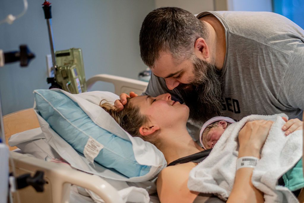 Man kissing a woman in a hospital bed while holding their newborn baby