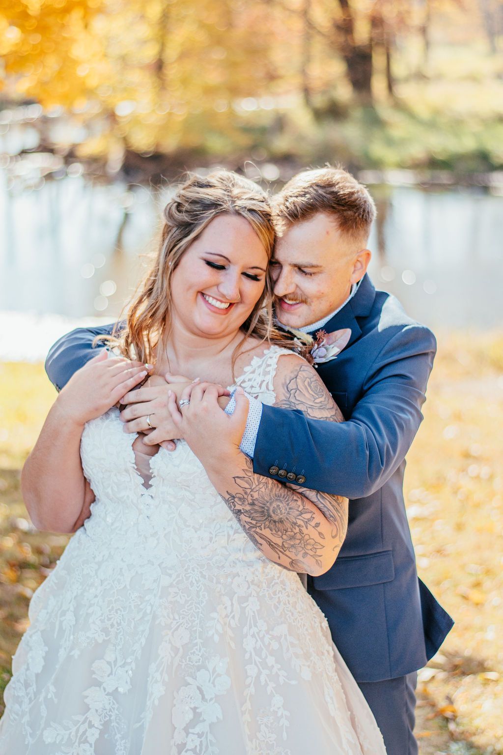 Couple embracing outdoors in a sunlit autumn setting, with a woman in a white lace dress and a man in a blue suit