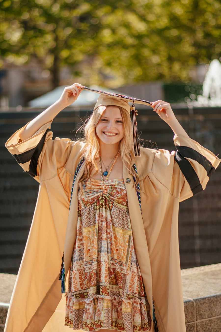 Smiling graduate in beige cap and gown holding mortarboard outdoors