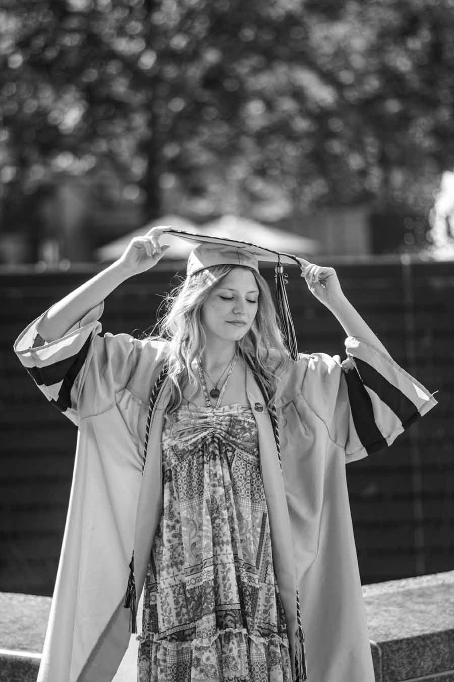 Person in a flowing robe holds a hat overhead outdoors, standing in front of a blurred fountain.