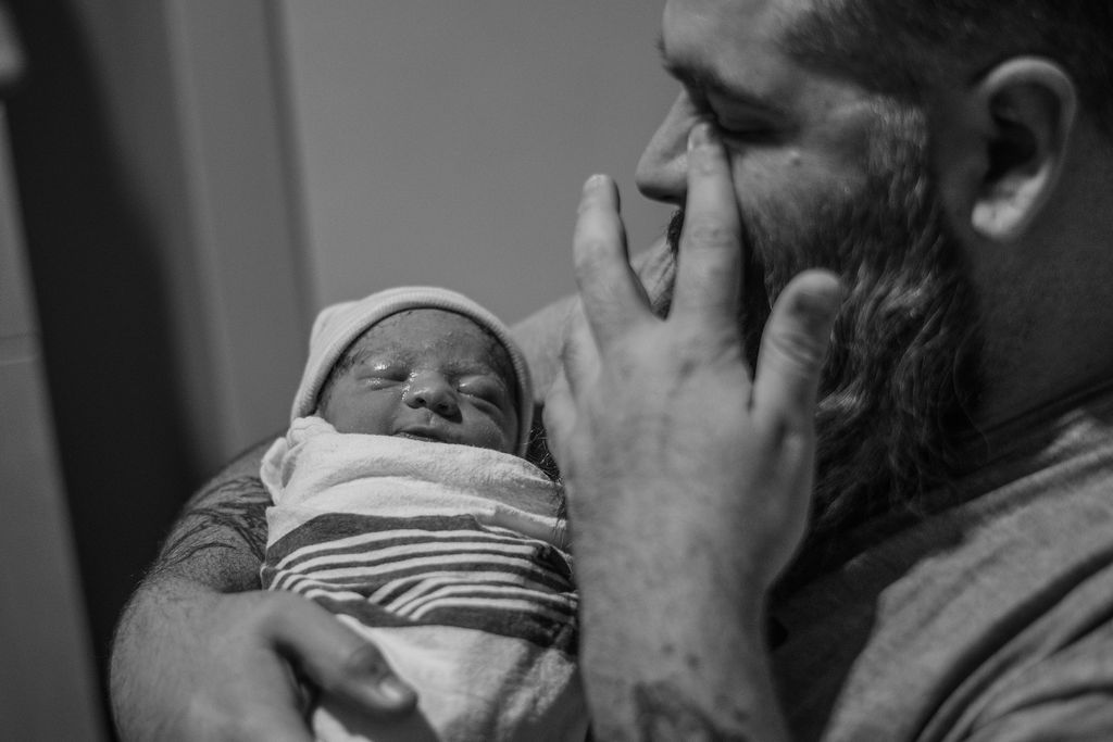 Man holding a sleeping newborn wrapped in a striped blanket, touching his face tenderly.