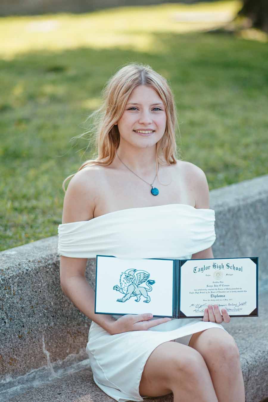 Woman in white dress sitting outdoors, holding an open certificate and smiling.