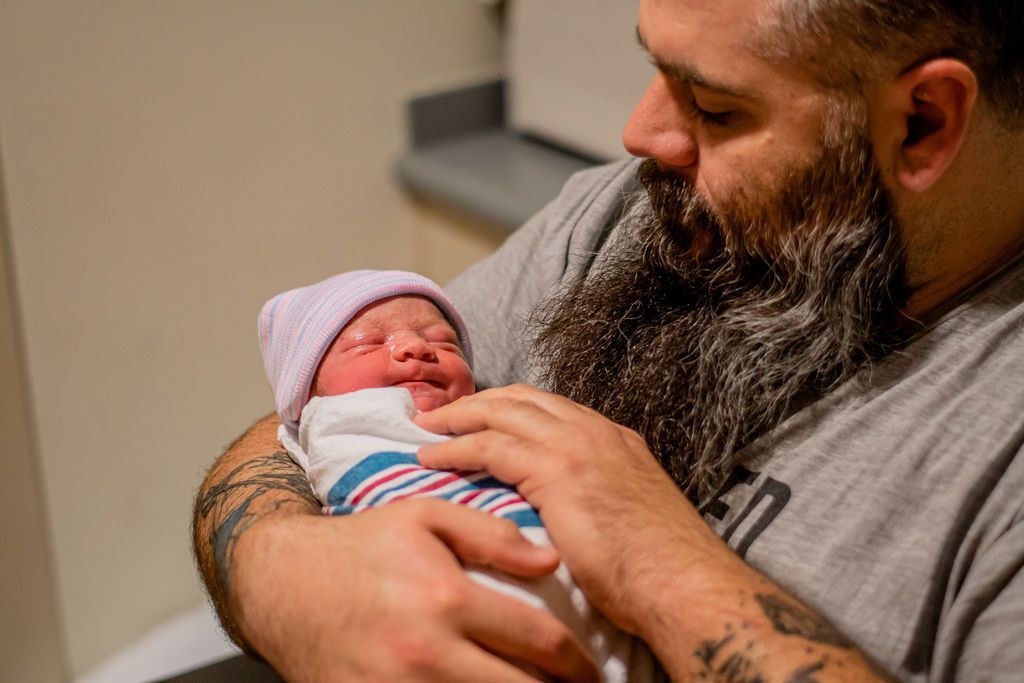 Man cradling a newborn wrapped in a striped blanket, gently touching the baby’s chest.
