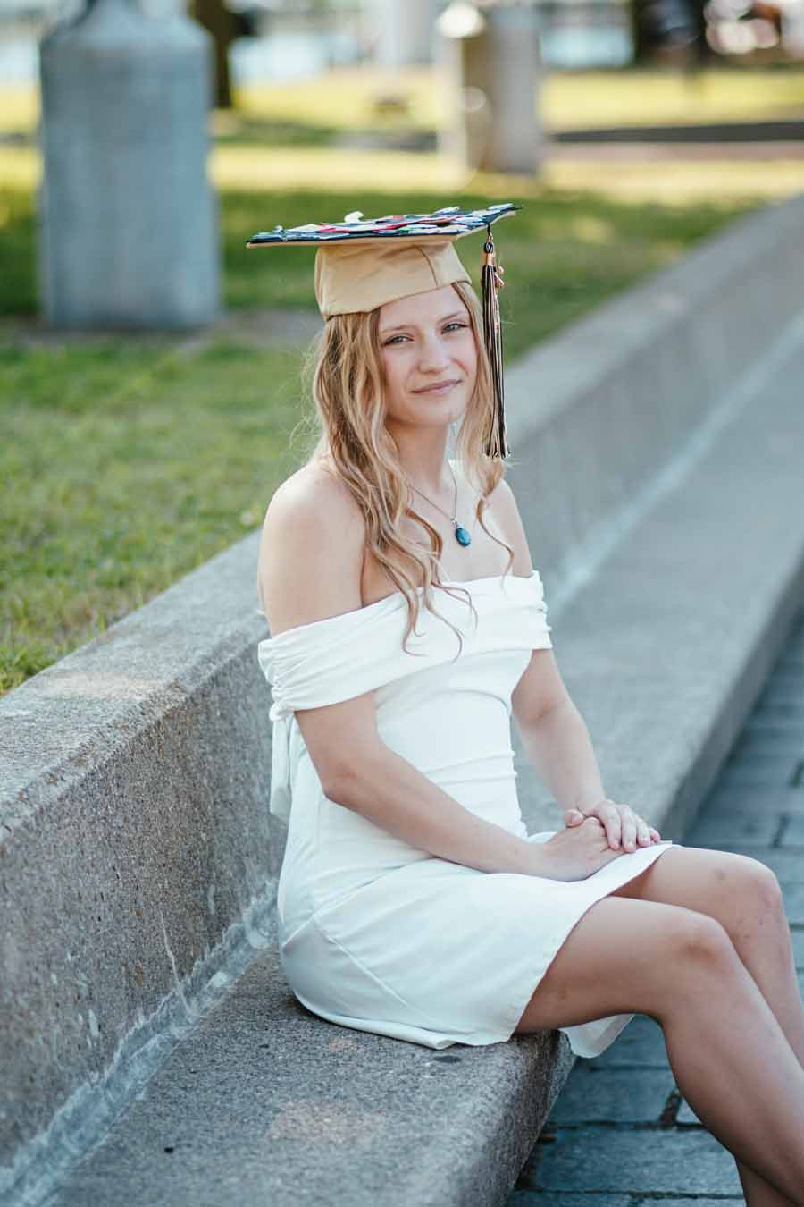 Woman in a white dress sitting on a curb, wearing a small graduation cap outdoors.