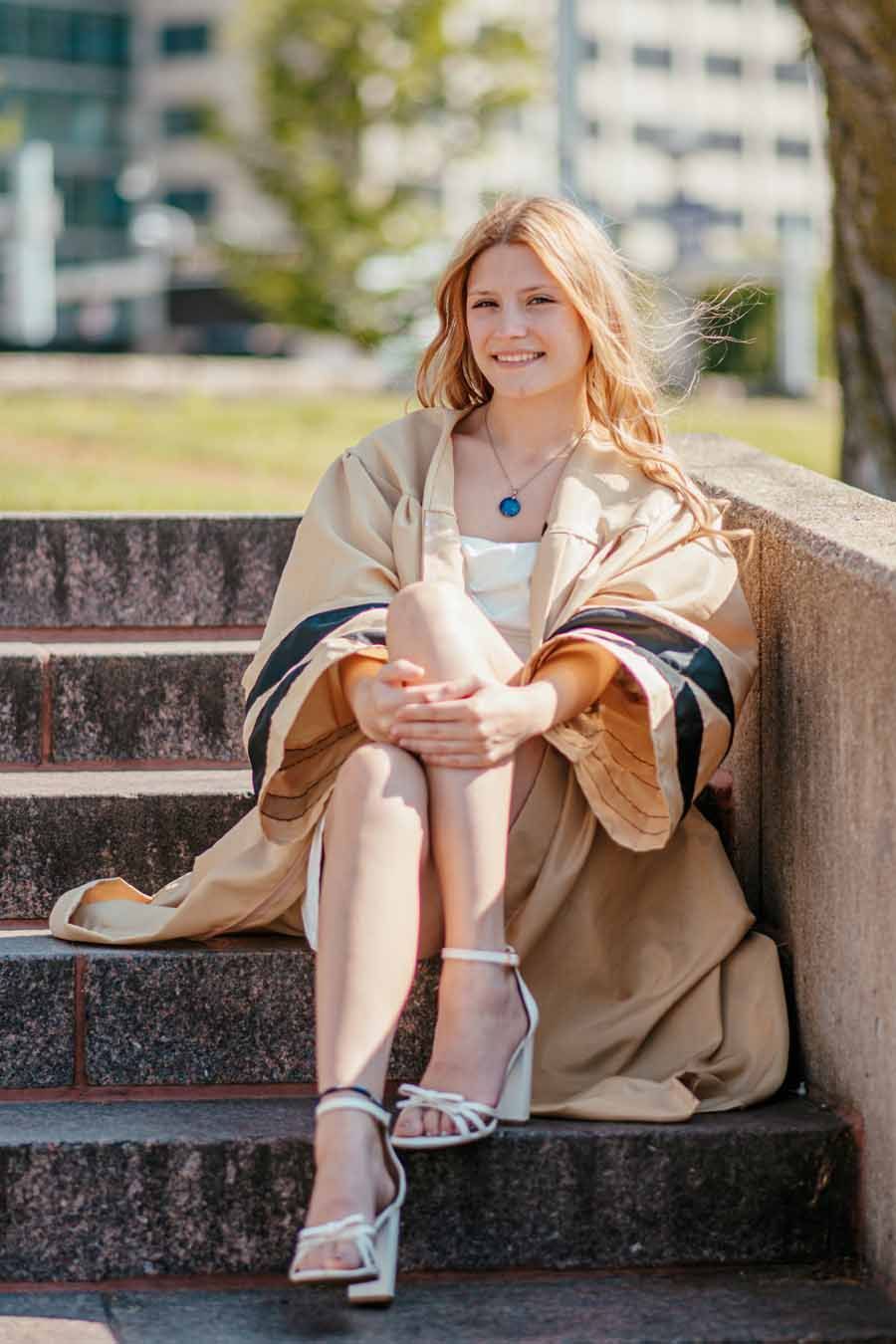 Woman sitting on steps outdoors, wearing a beige shawl and sandals, smiling in sunlight.