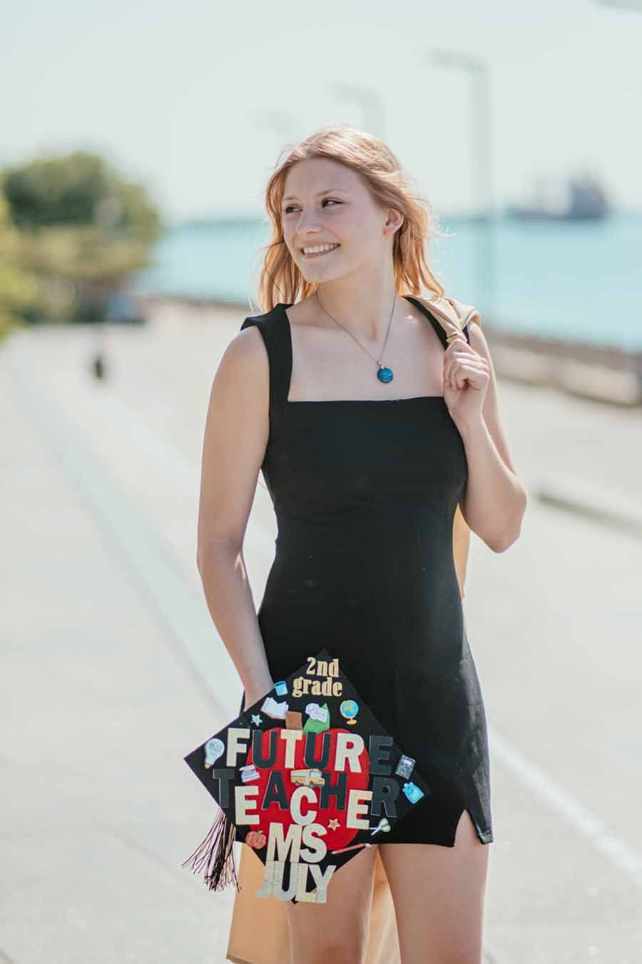 Woman in a black dress smiling on a sunny waterfront path, holding a patterned bag