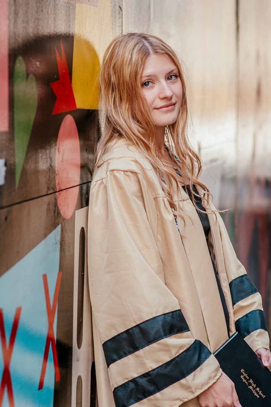 Young woman in a beige coat with black stripes stands beside a colorful mural, looking at the camera.