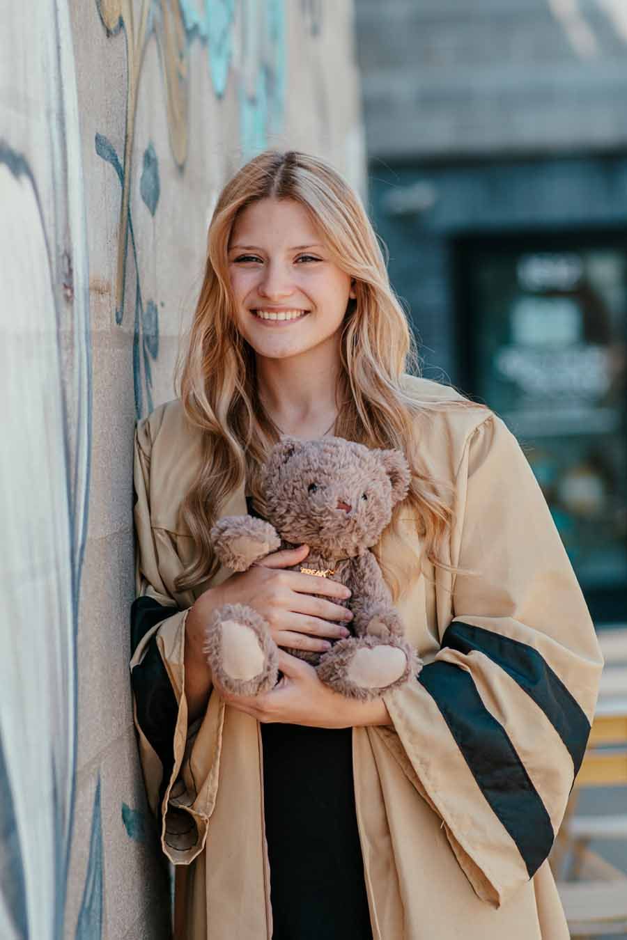 Smiling person holding a teddy bear beside a mural wall outdoors