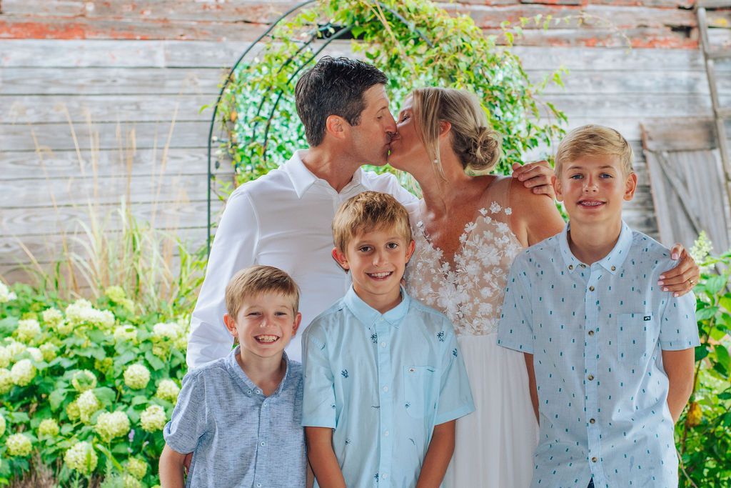 Family of five outdoors, parents kissing while four children in light blue shirts stand in front of a white fence.