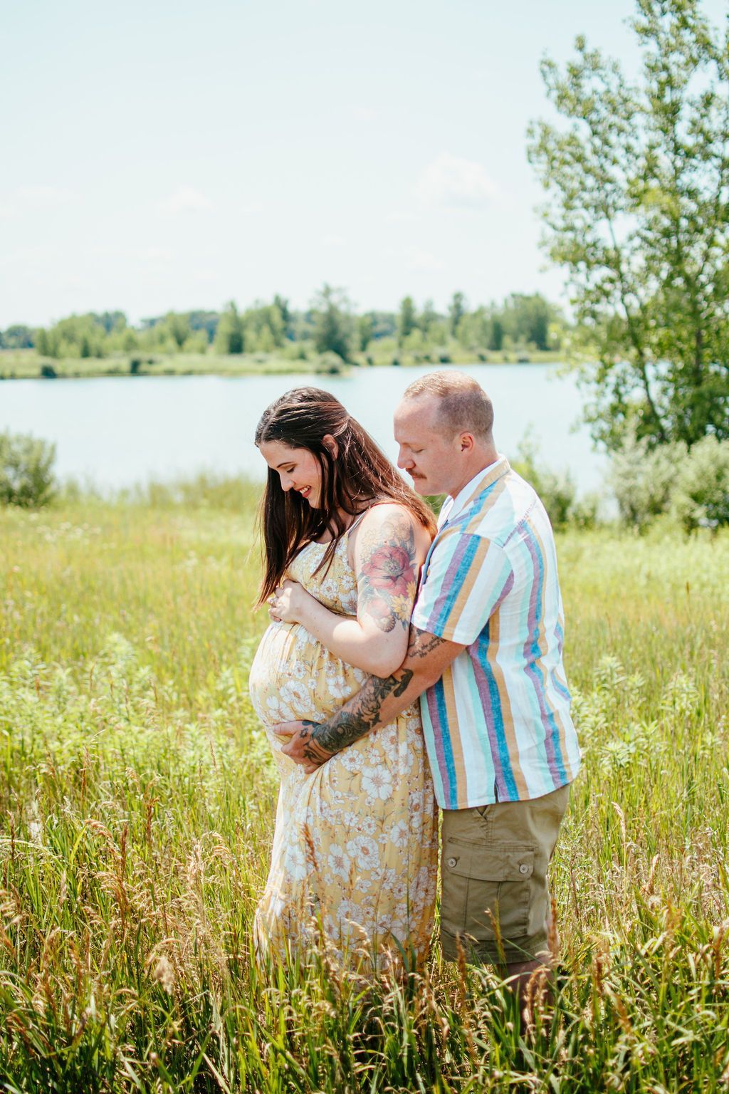 Couple embracing in a grassy field by a lake, with the woman visibly pregnant.