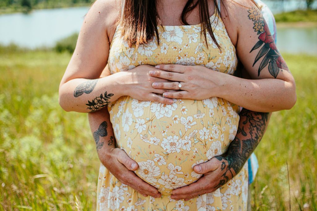 Two people in a field gently hold a pregnant belly, with tattoos visible and a yellow dress centered.