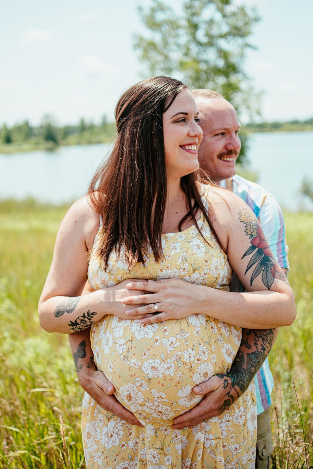 Couple embracing in a grassy field by a lake, smiling under a bright sky.