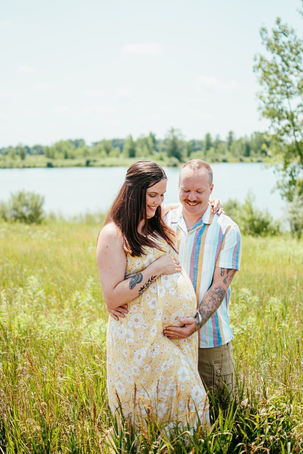 Two people standing in tall grass by a lake, smiling and embracing a pregnant belly