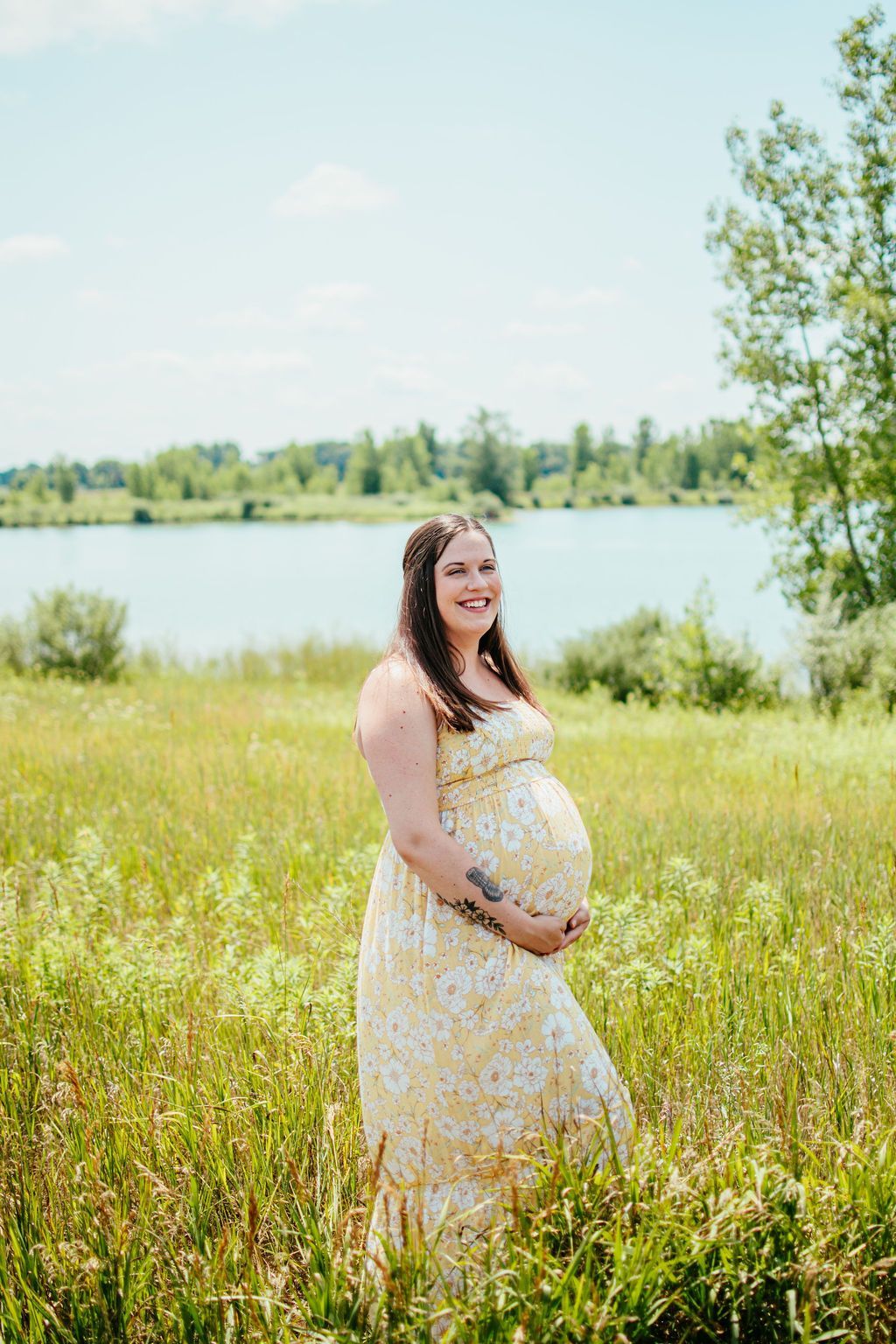 Pregnant woman in a light floral dress standing in a grassy field by a lake, smiling.