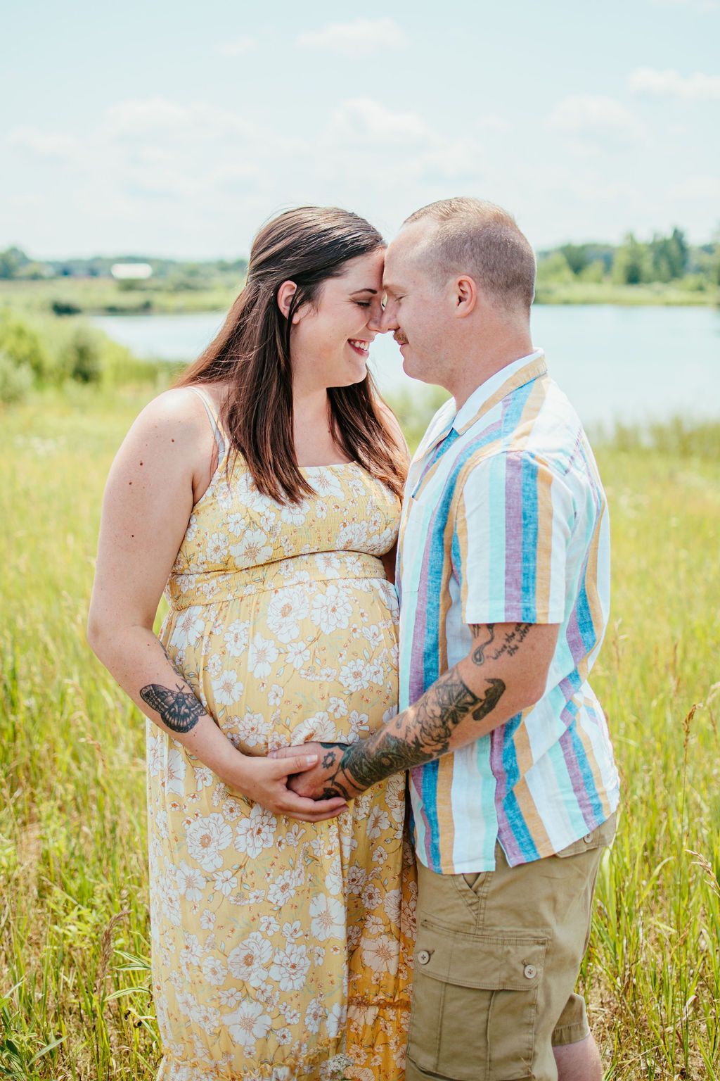 Pregnant couple standing close by a lake, touching foreheads in a sunny field.