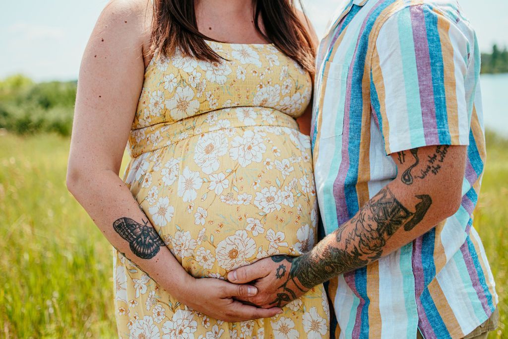 Couple standing outdoors, hands clasped over a pregnant belly, wearing tattoos and colorful clothing