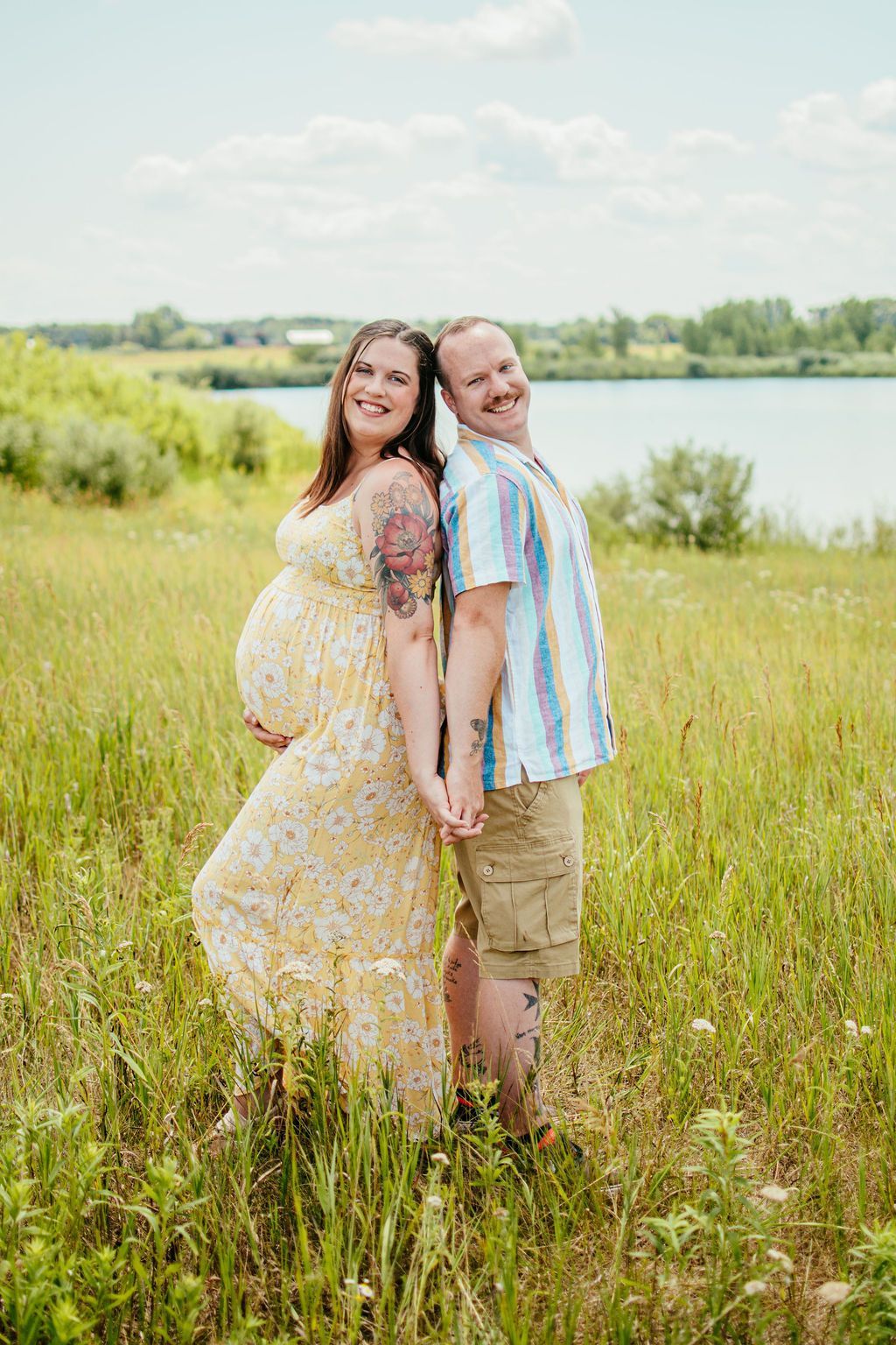 Two people standing back-to-back in a grassy field by a lake, smiling under a bright sky.