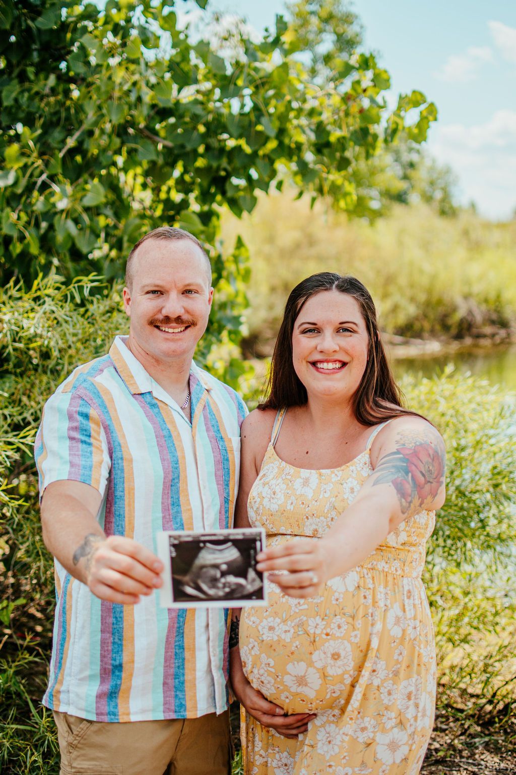 Couple holding an ultrasound photo outdoors, smiling in a leafy setting, with the woman pregnant.