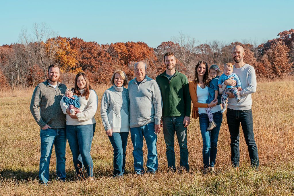 Eight people standing together in a grassy field with autumn trees in the background