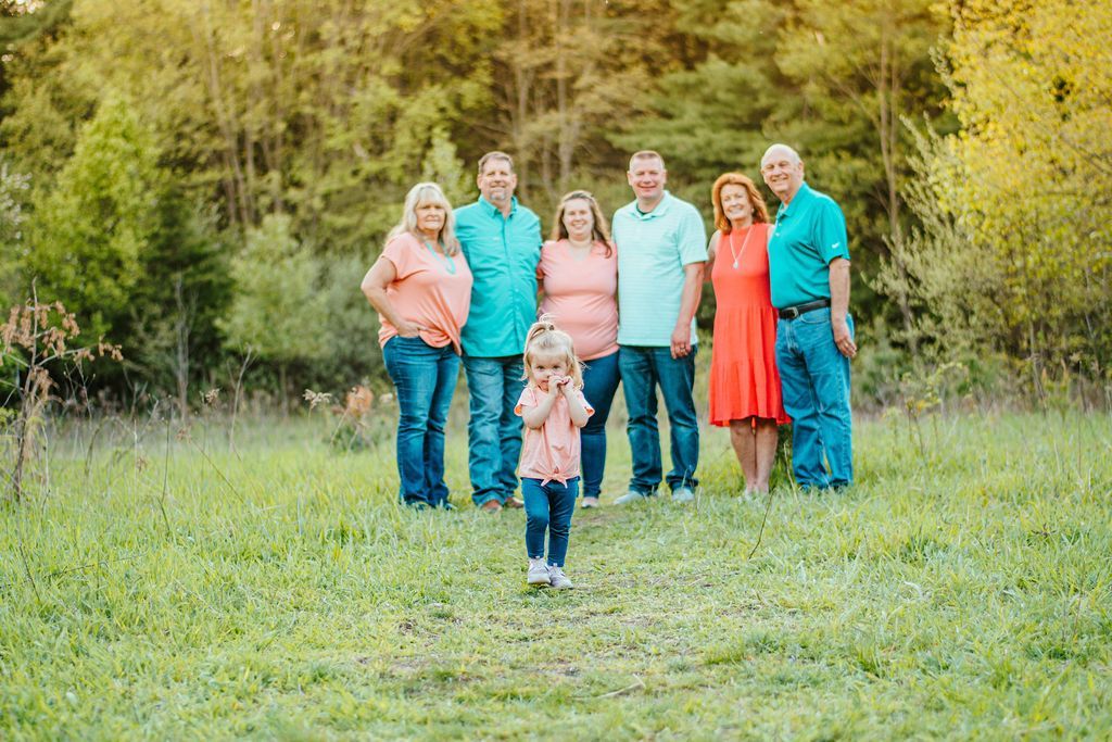 Family standing in a grassy field, with a child walking toward the camera and trees in the background