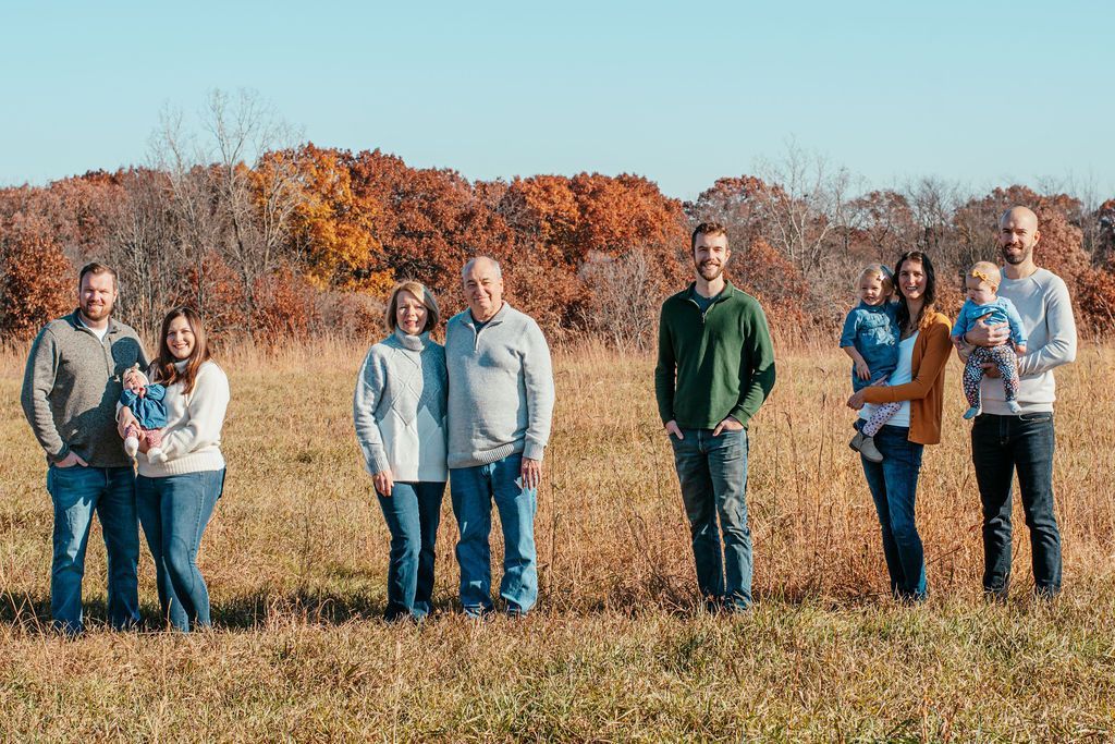 Seven people standing in a grassy field with autumn trees in the background
