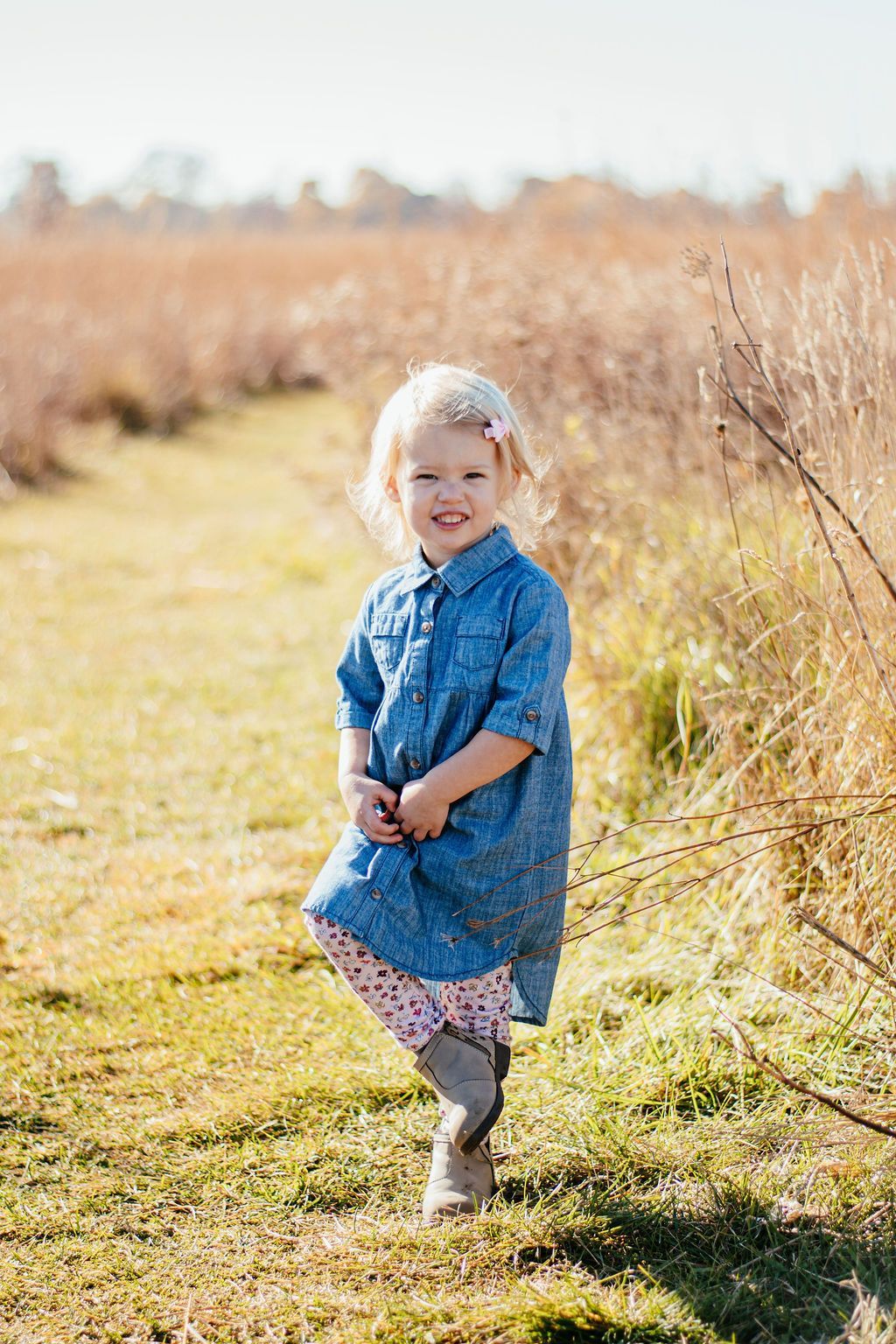 Child in a blue dress standing on a sunlit path beside tall grass, smiling at the camera.
