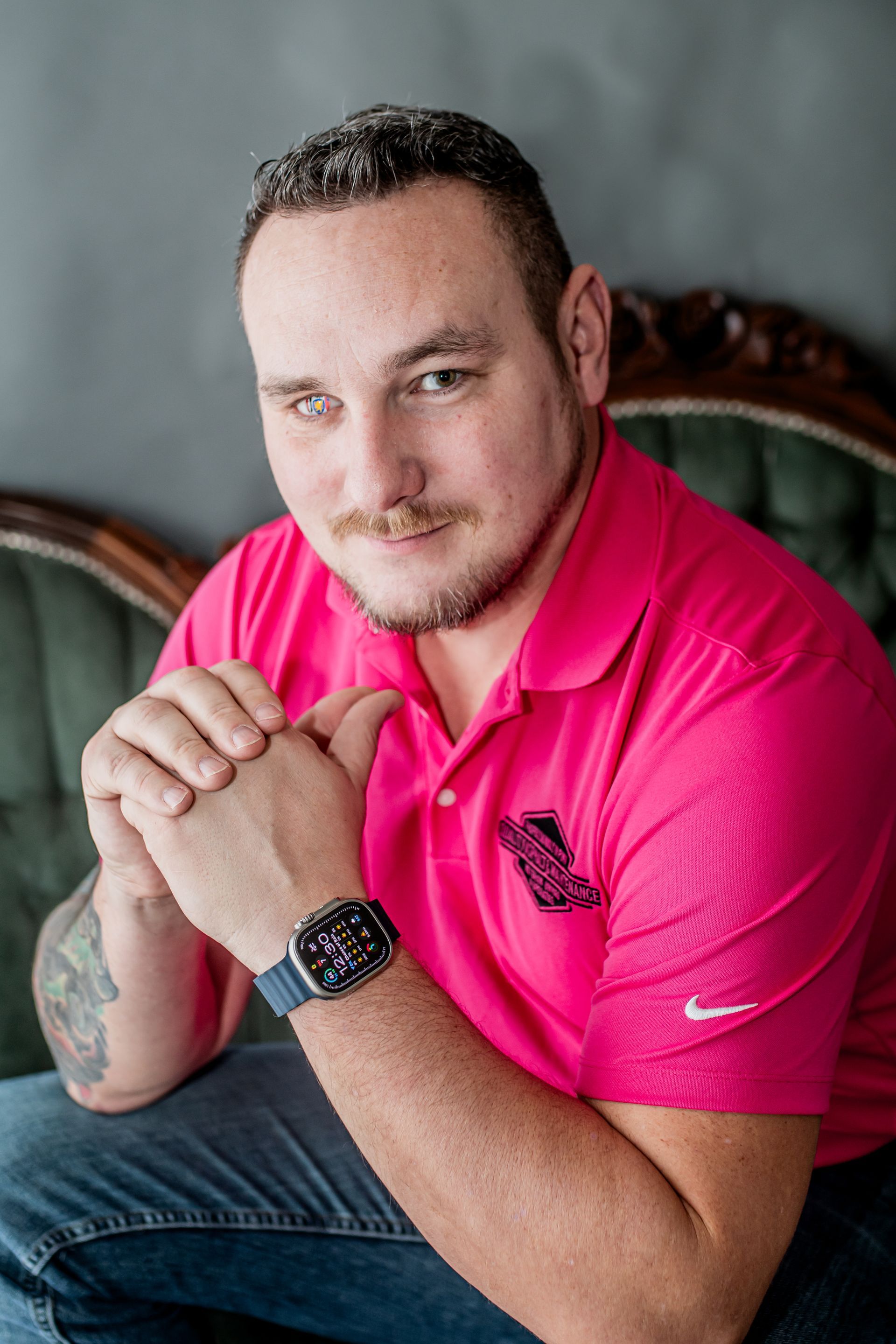 Man in a bright pink polo shirt seated with clasped hands, smiling against a gray backdrop.