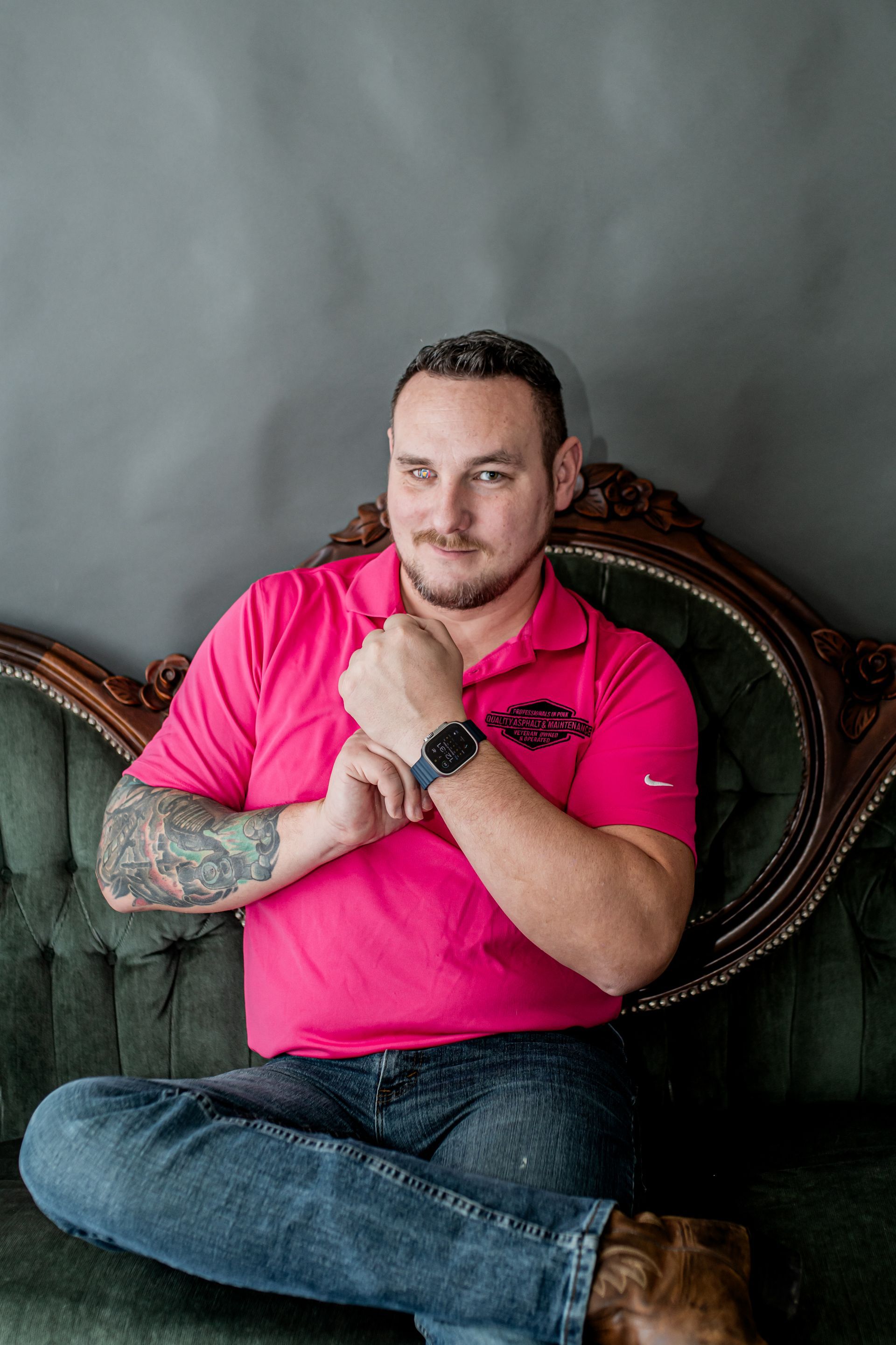 Man in bright pink shirt sitting on a green sofa with arms crossed against a gray wall