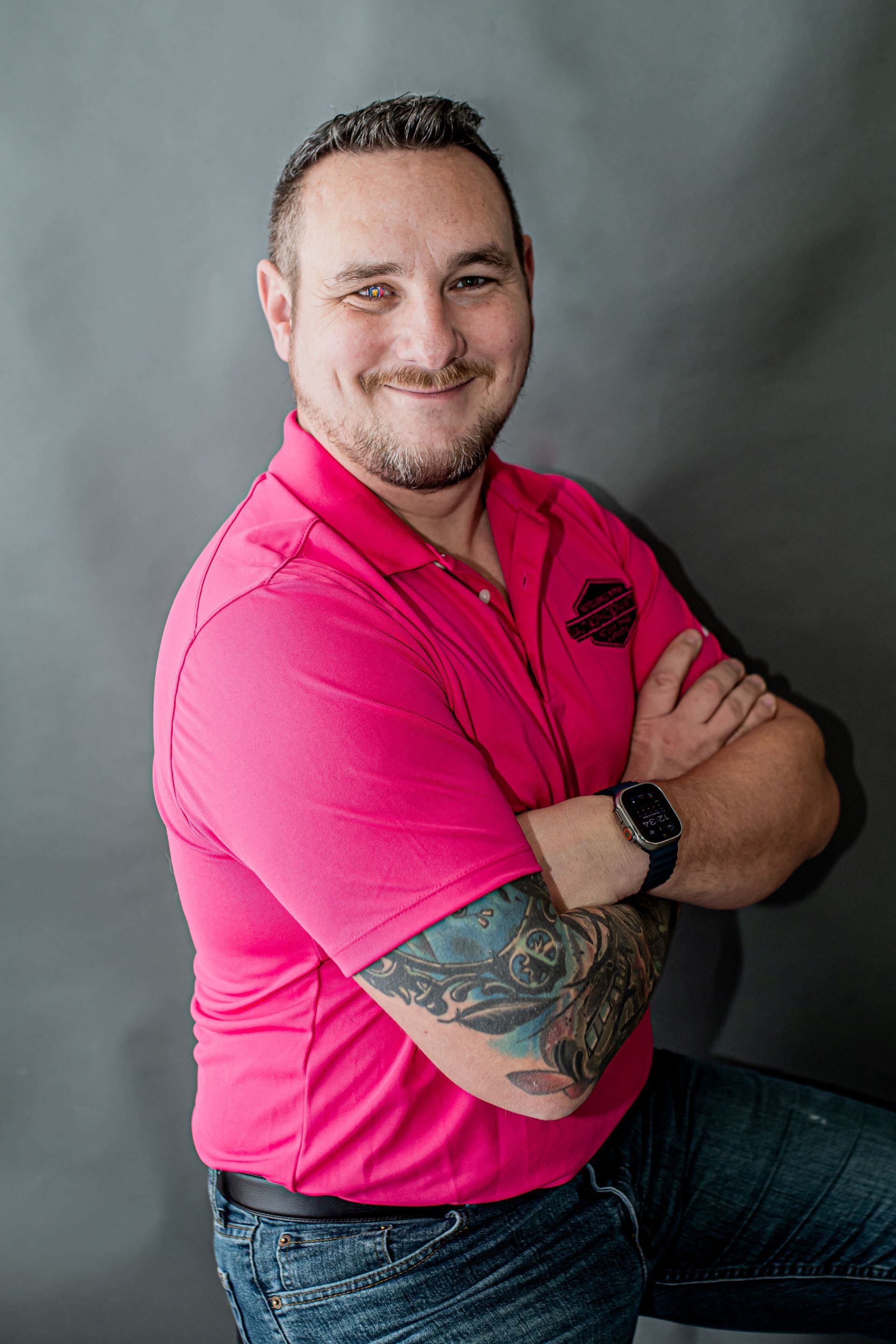 Man in bright pink polo shirt with tattooed arm, smiling with arms crossed against a gray backdrop