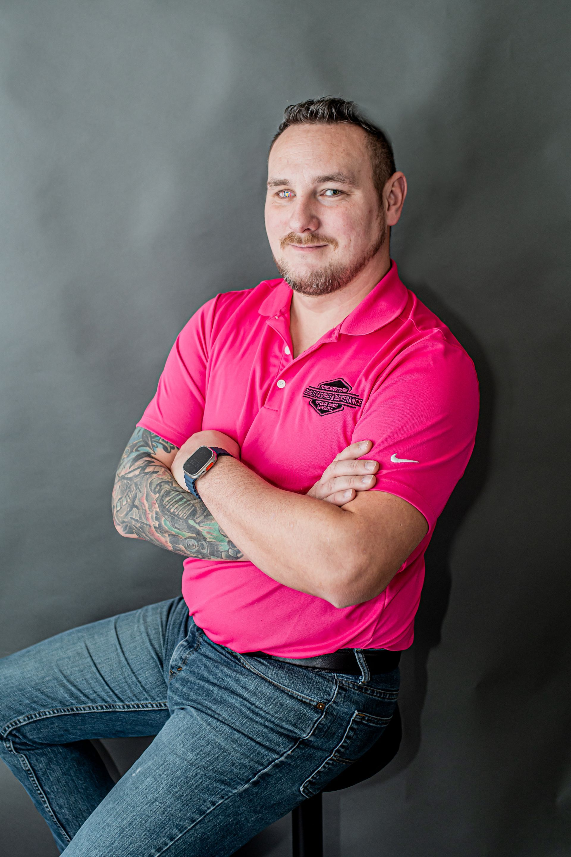 Man seated with arms crossed, wearing a bright pink polo and jeans against a gray backdrop.
