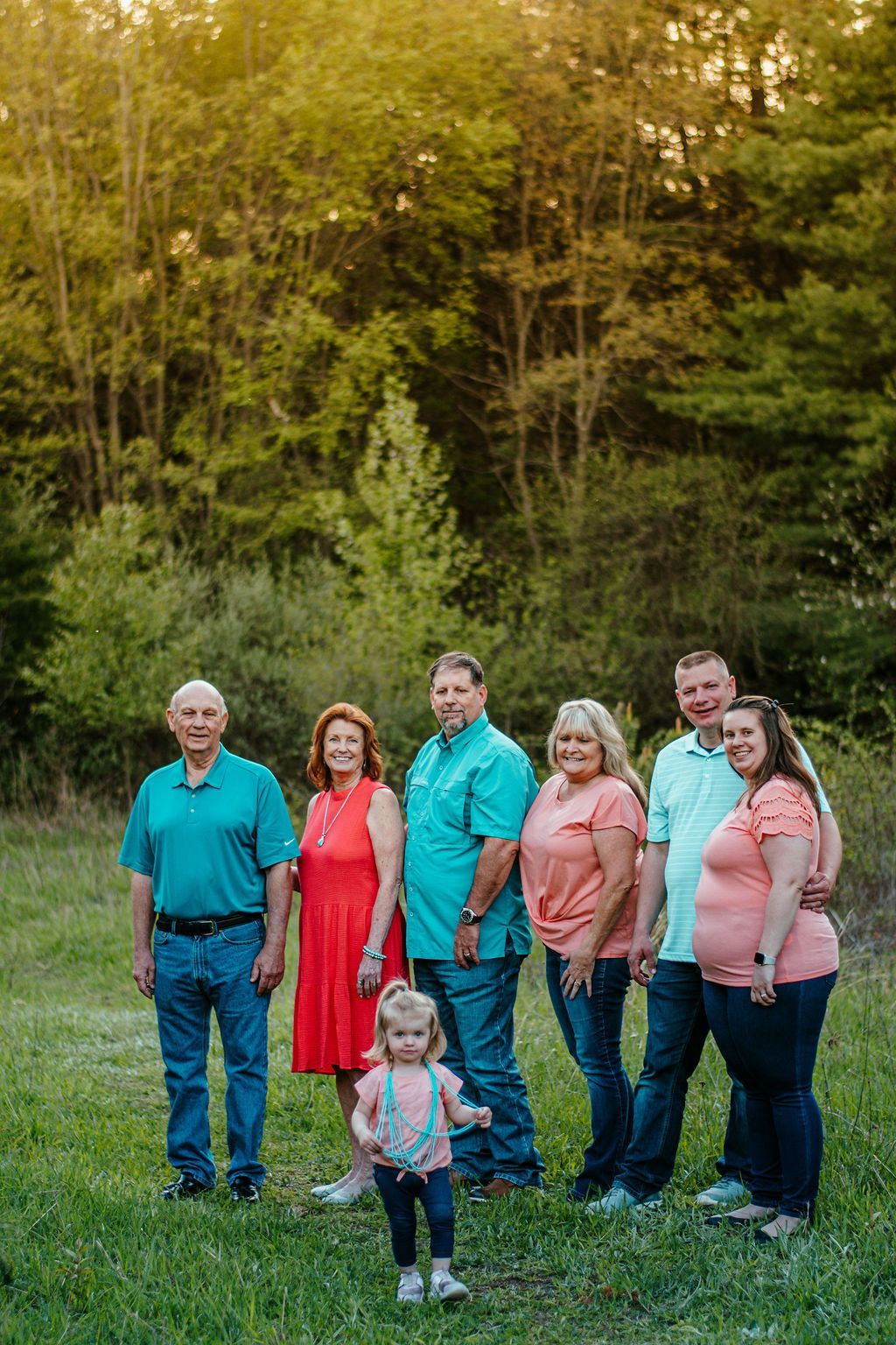 Group of seven standing in a grassy field at the edge of a wooded area, with a child in front