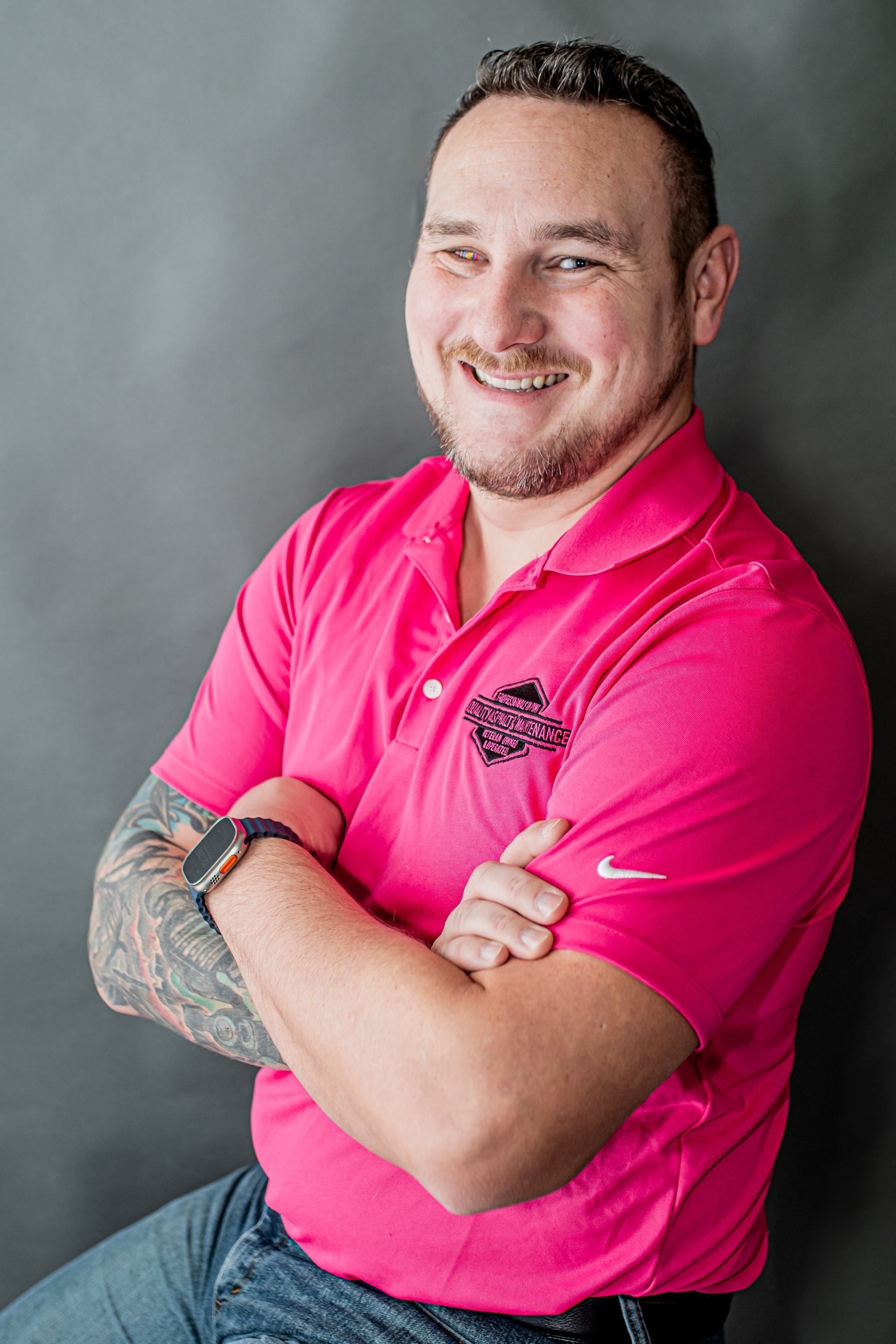 Smiling man in a bright pink polo shirt with tattooed arm, seated with arms crossed against a gray backdrop