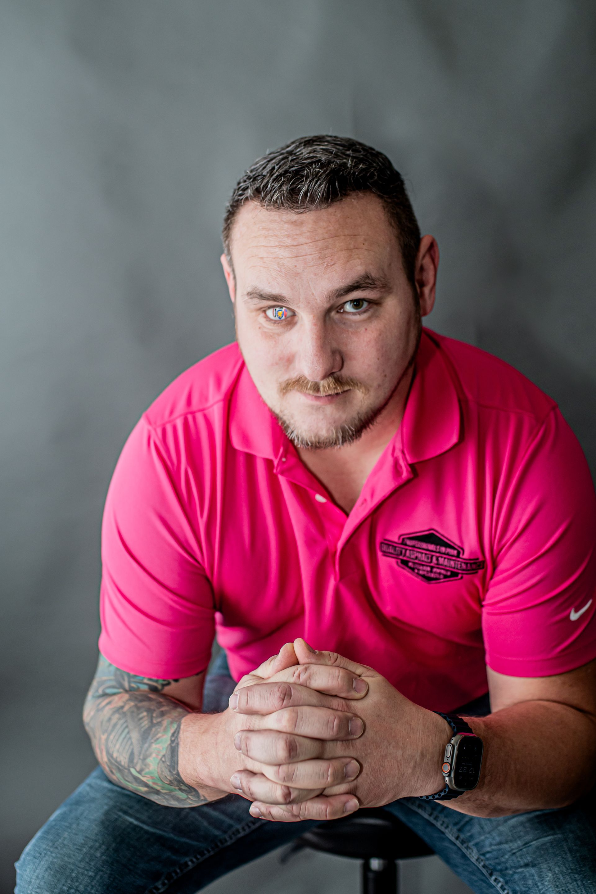 Man in a bright pink polo shirt seated with hands clasped against a gray studio backdrop
