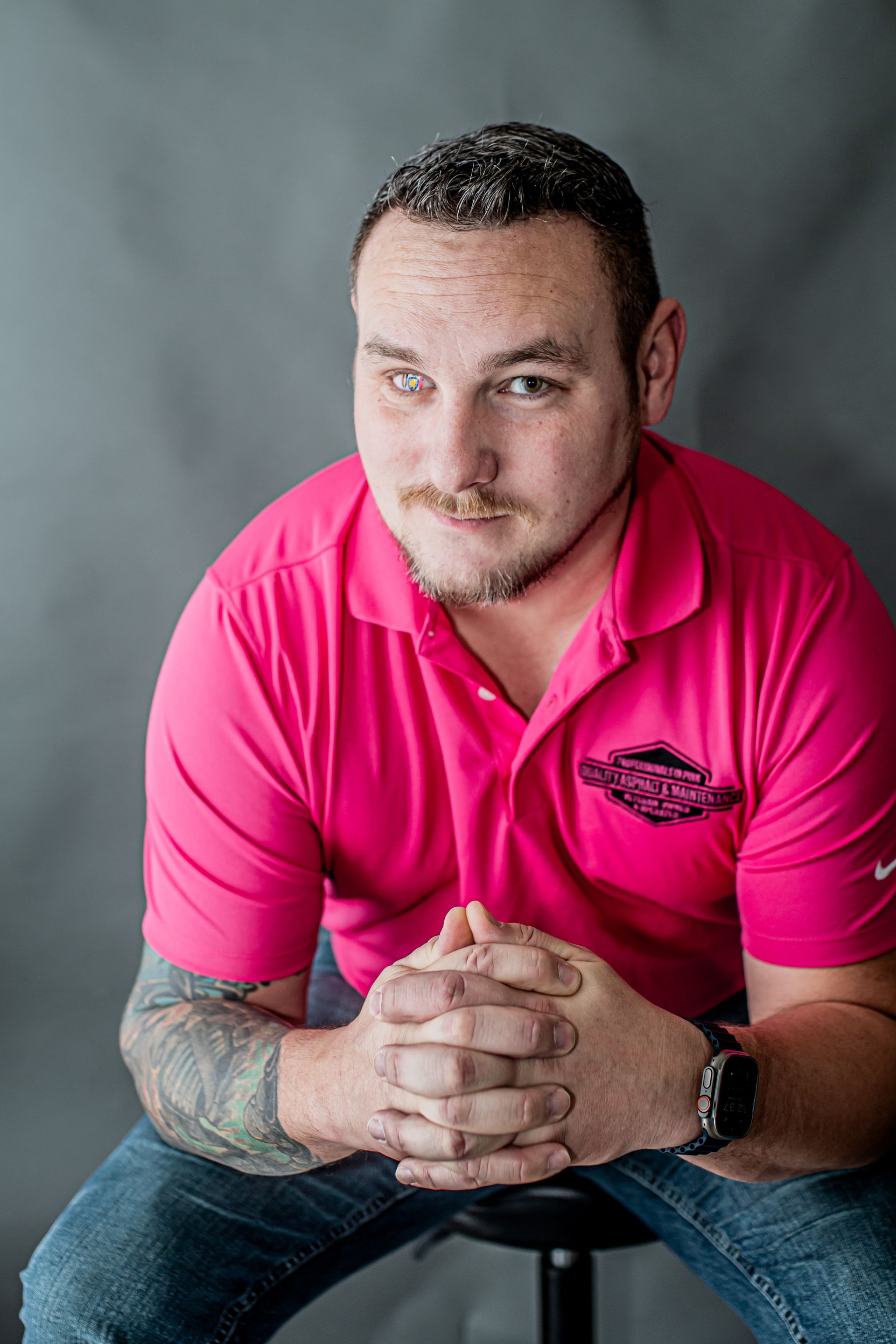 Man seated against a gray backdrop, wearing a bright pink polo shirt, hands clasped