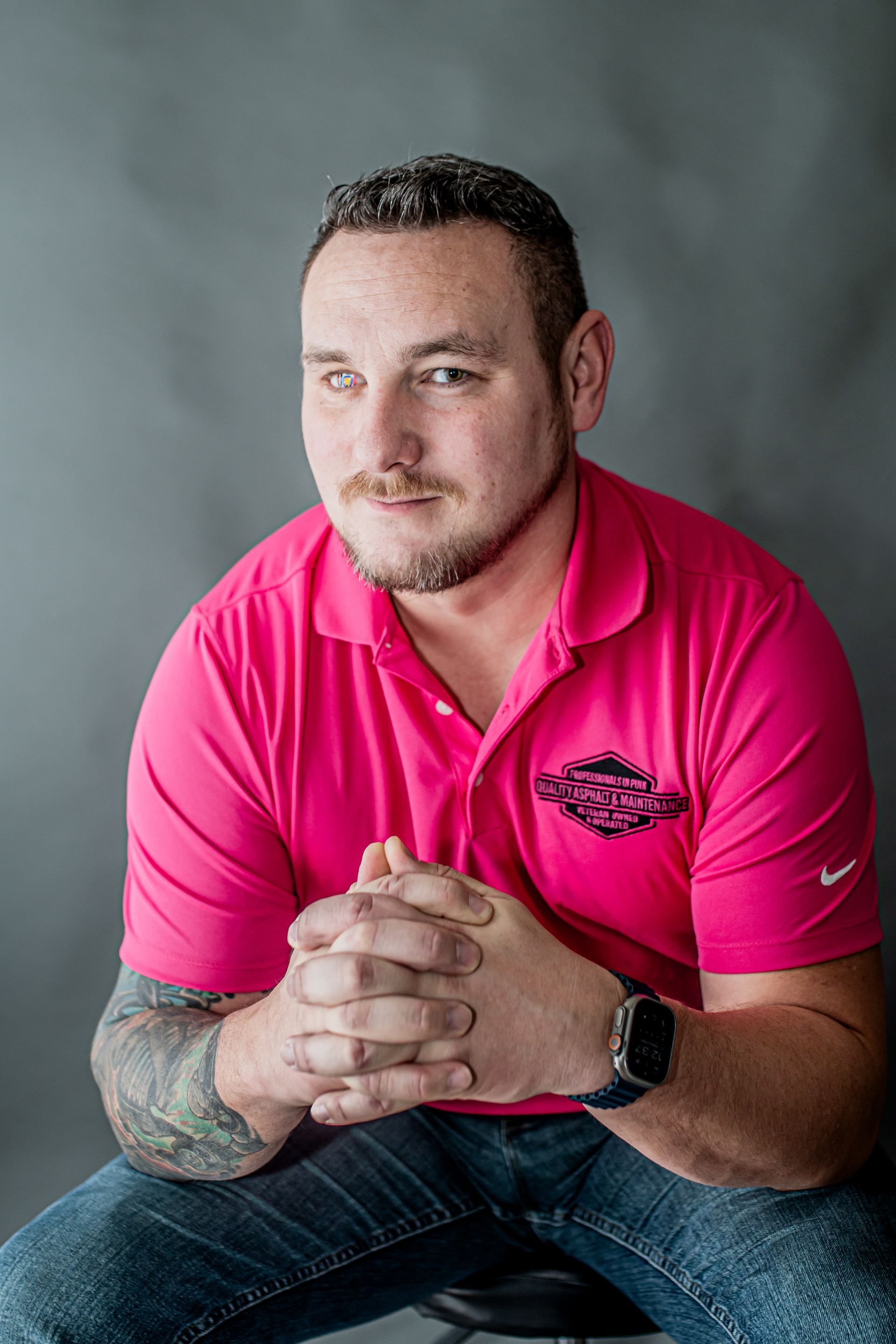 Man seated in a bright pink polo shirt, hands clasped, against a gray studio backdrop.