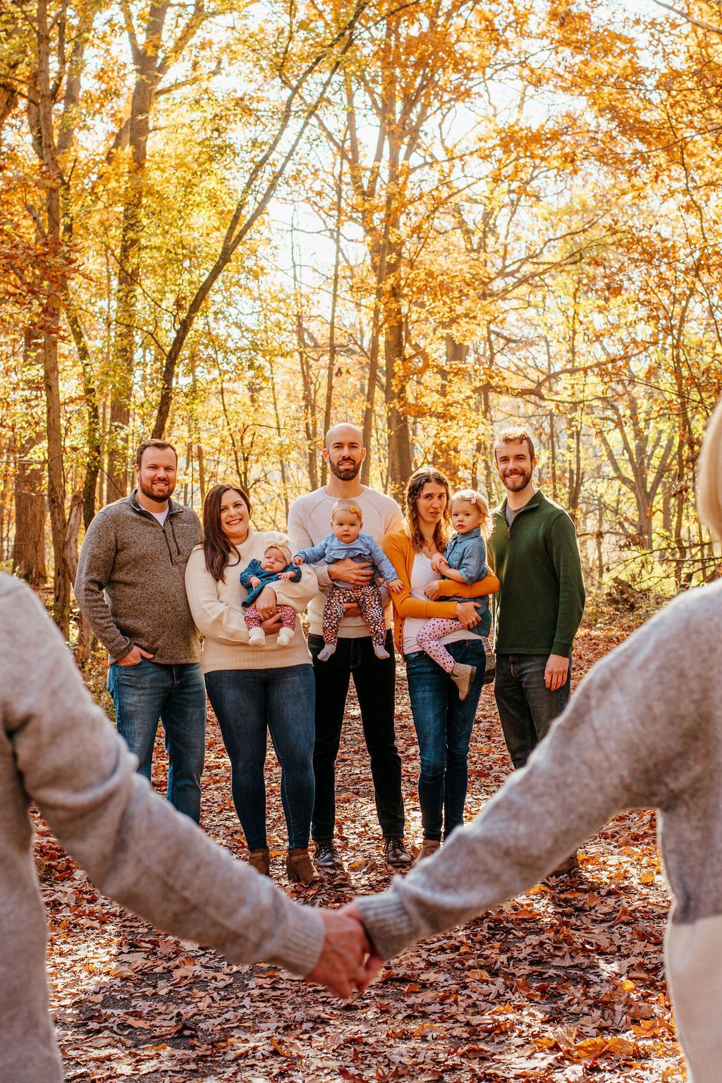 Five people standing in a wooded autumn setting, holding hands in a circle with a baby in the center.