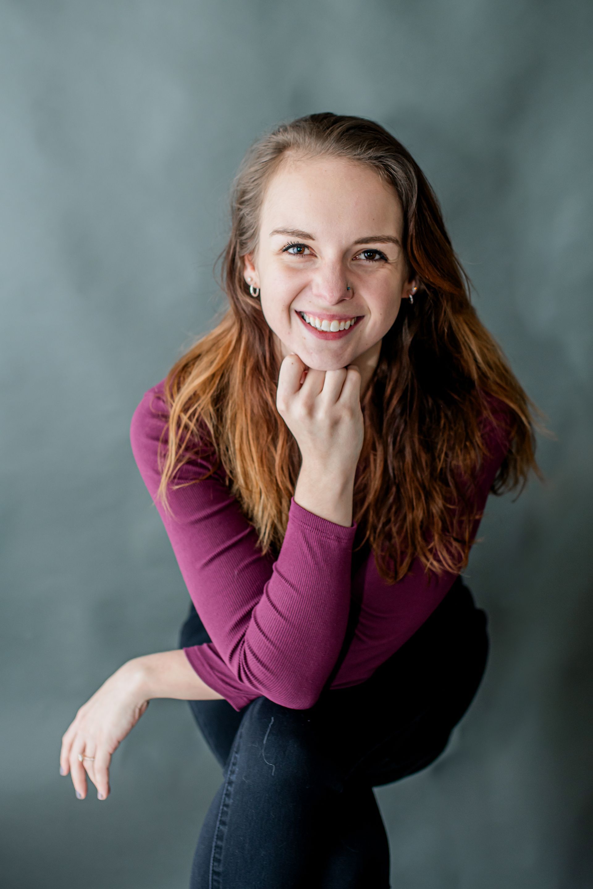 Smiling woman in a purple top and jeans, seated against a gray studio backdrop with one hand under her chin