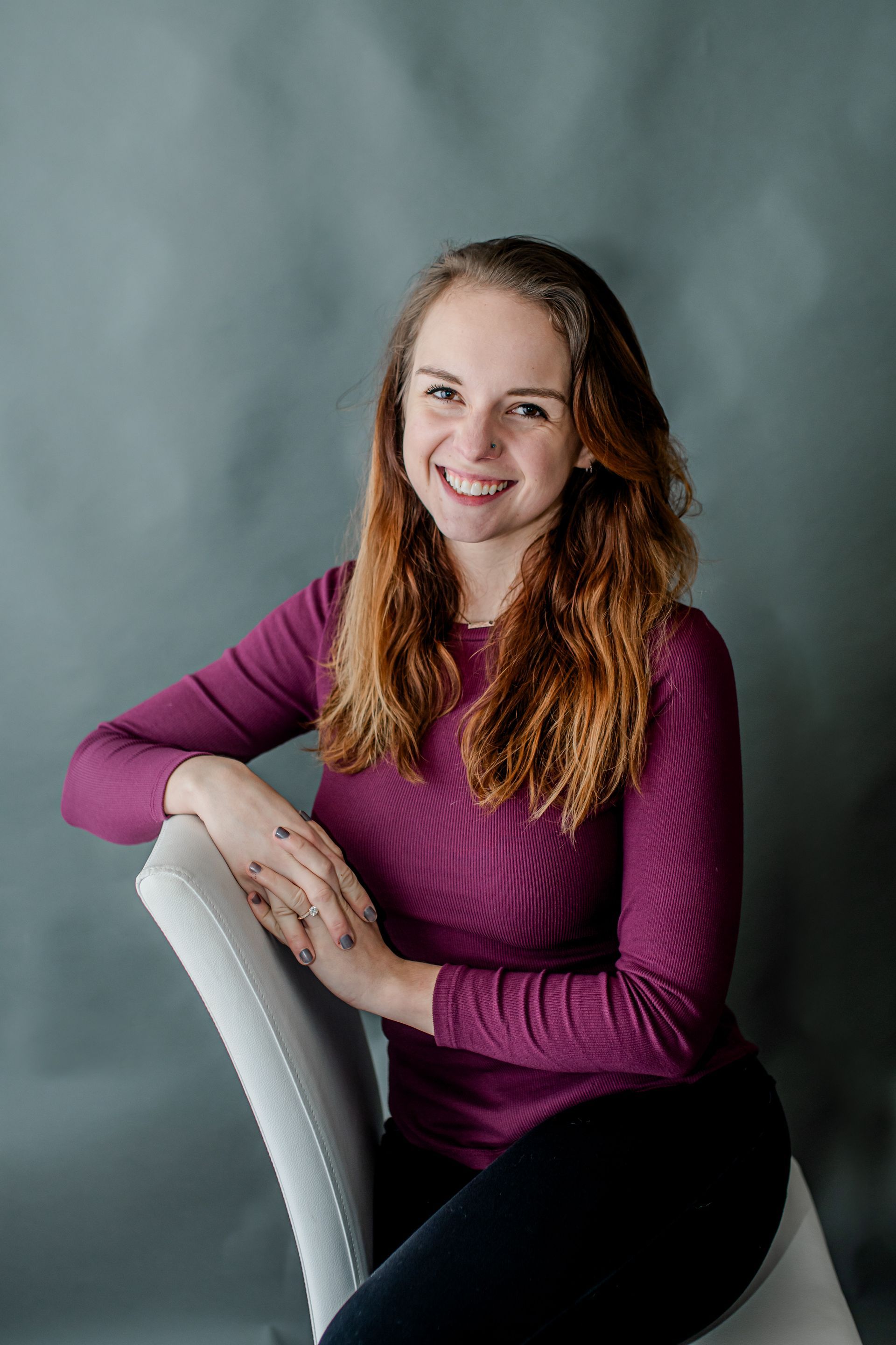 Smiling person in a purple sweater leaning on a white chair against a gray backdrop