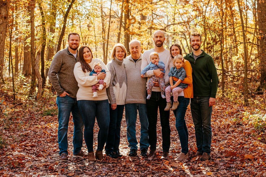 Family of eight standing together in a sunlit autumn forest, smiling at the camera.