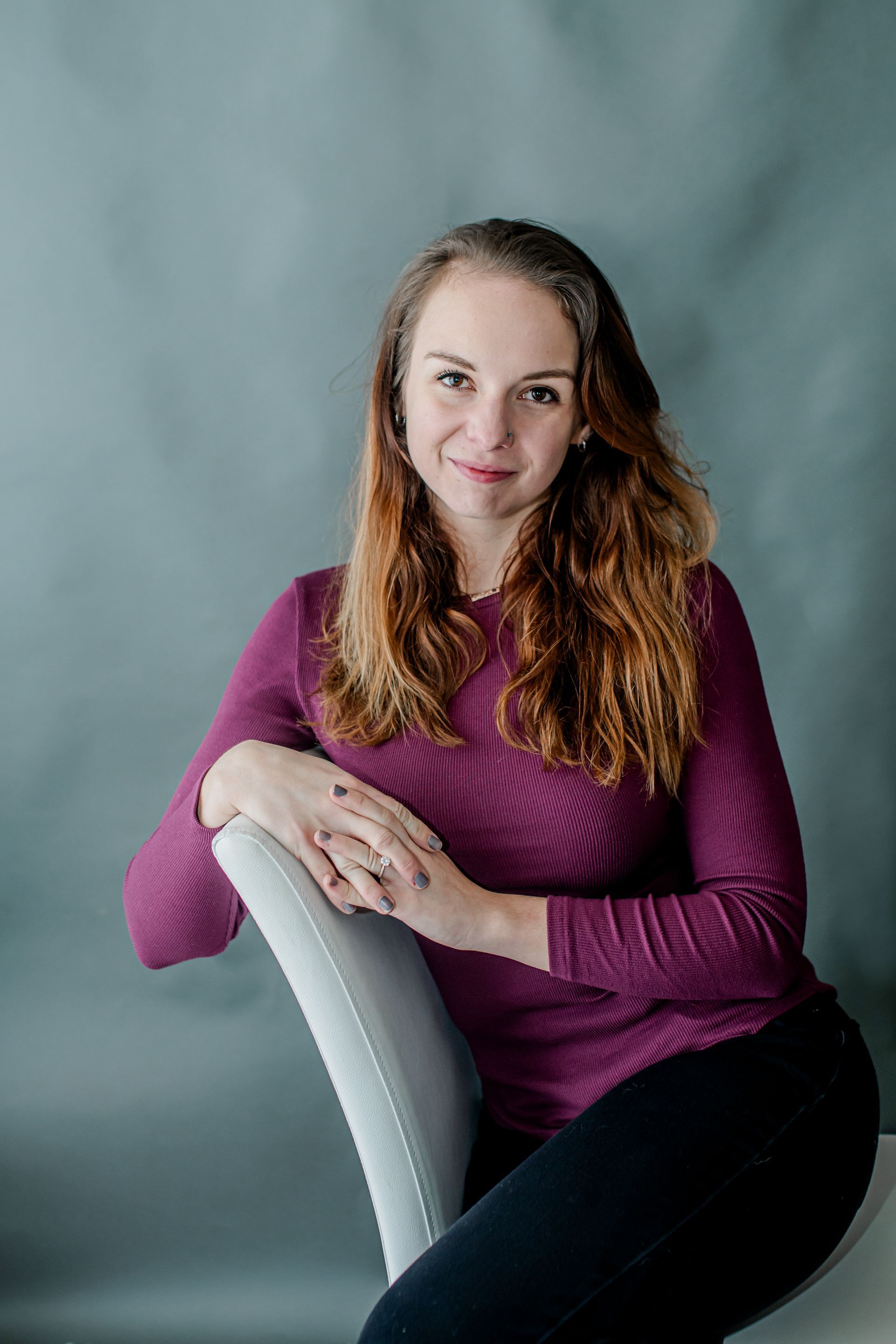 Woman in a purple sweater leaning on a white chair against a gray backdrop.