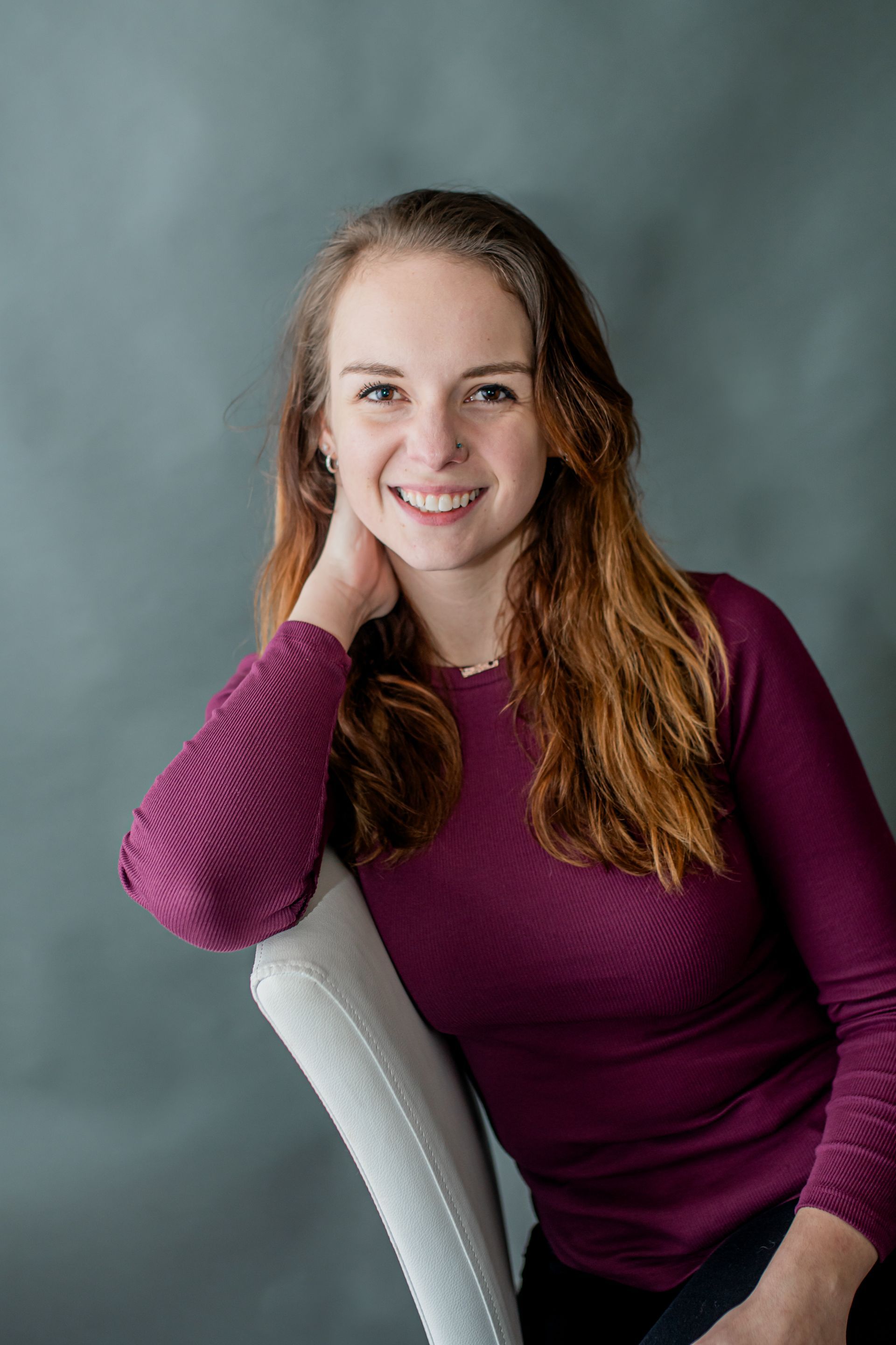 Smiling person with long auburn hair, wearing a purple top, leaning on a white chair against a gray backdrop