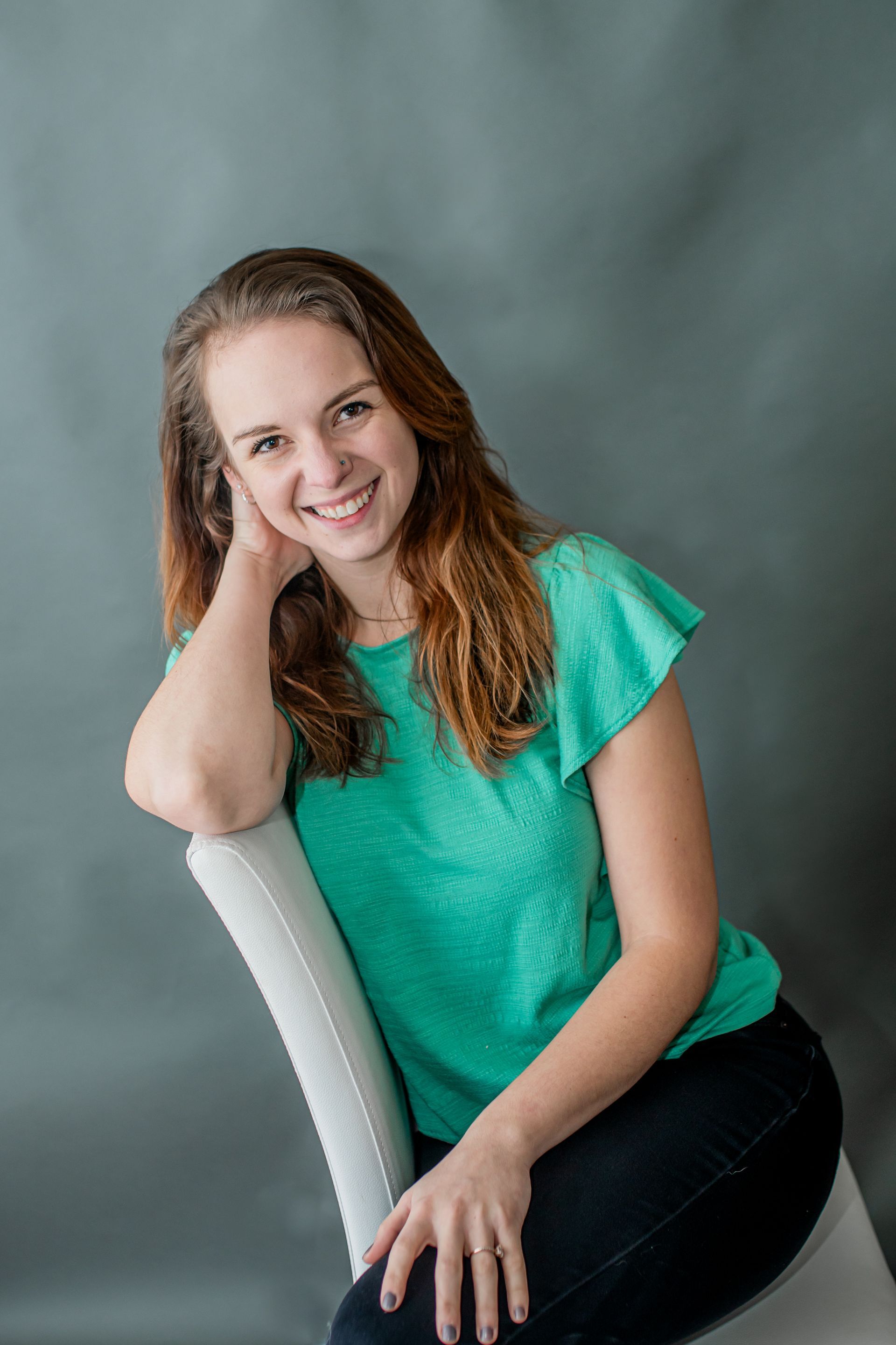 Smiling woman in teal top seated on a white chair against a gray backdrop