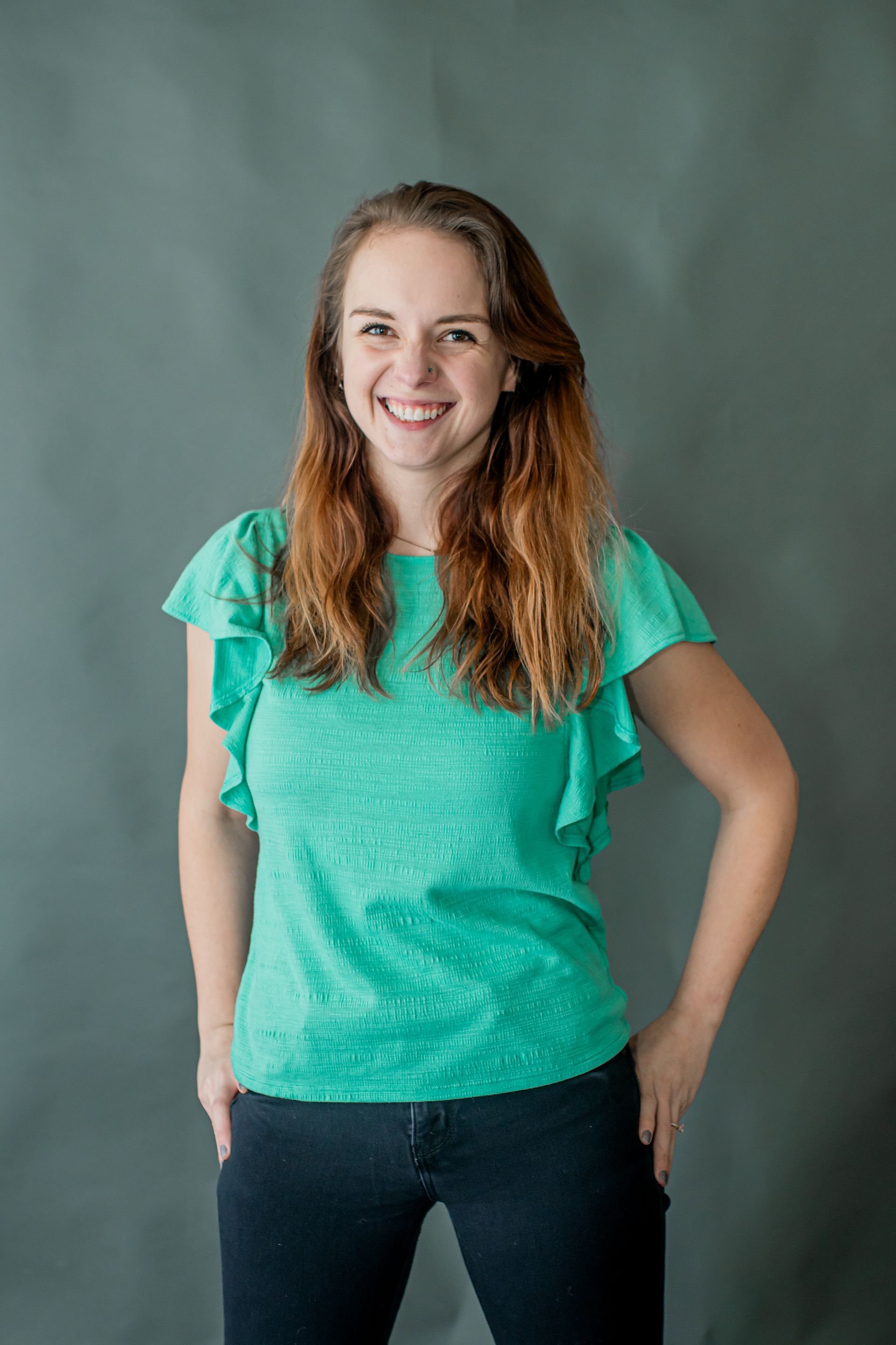 Smiling person in a teal shirt and jeans standing against a gray backdrop