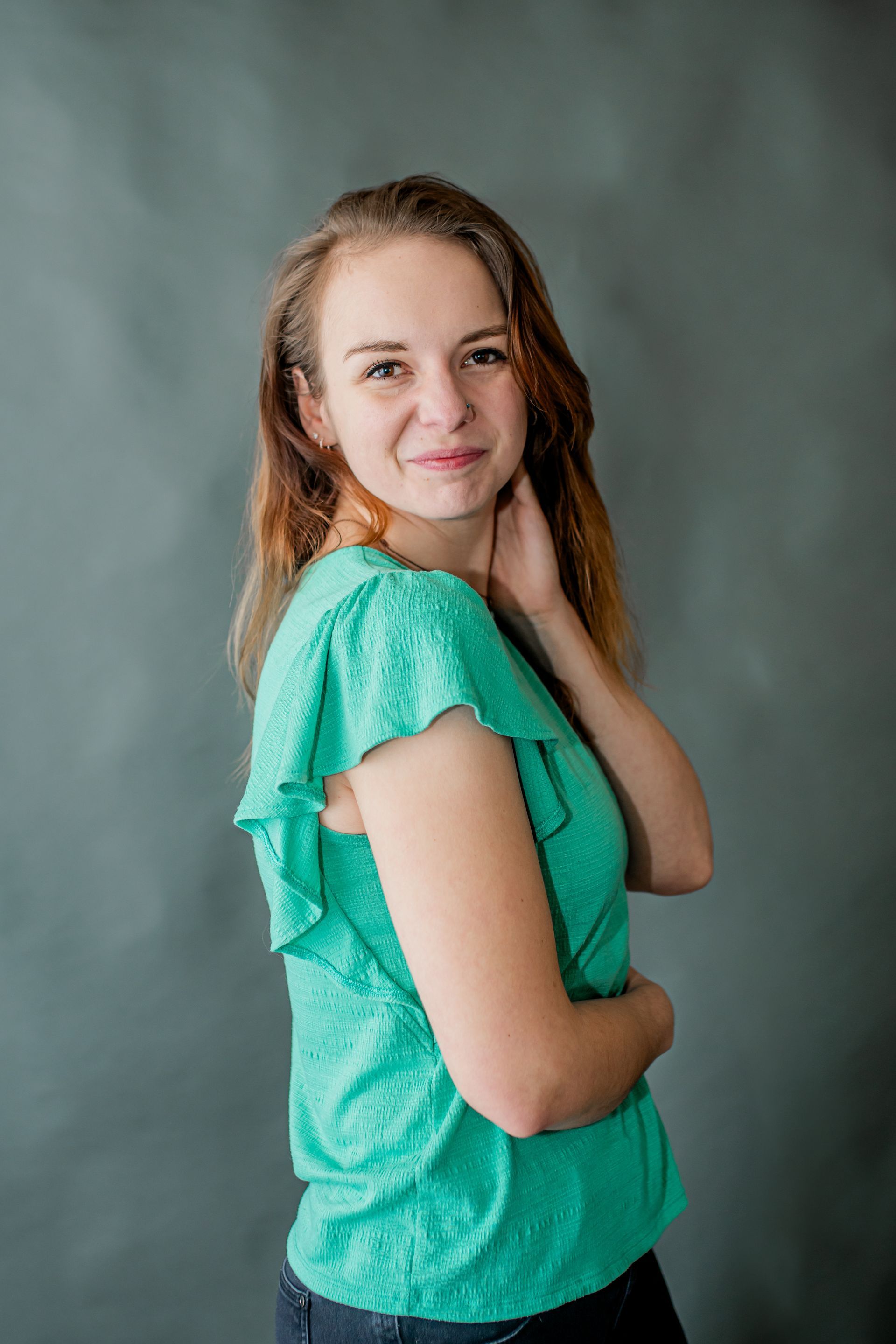 Woman in a turquoise top posing with hand on chin against a gray backdrop