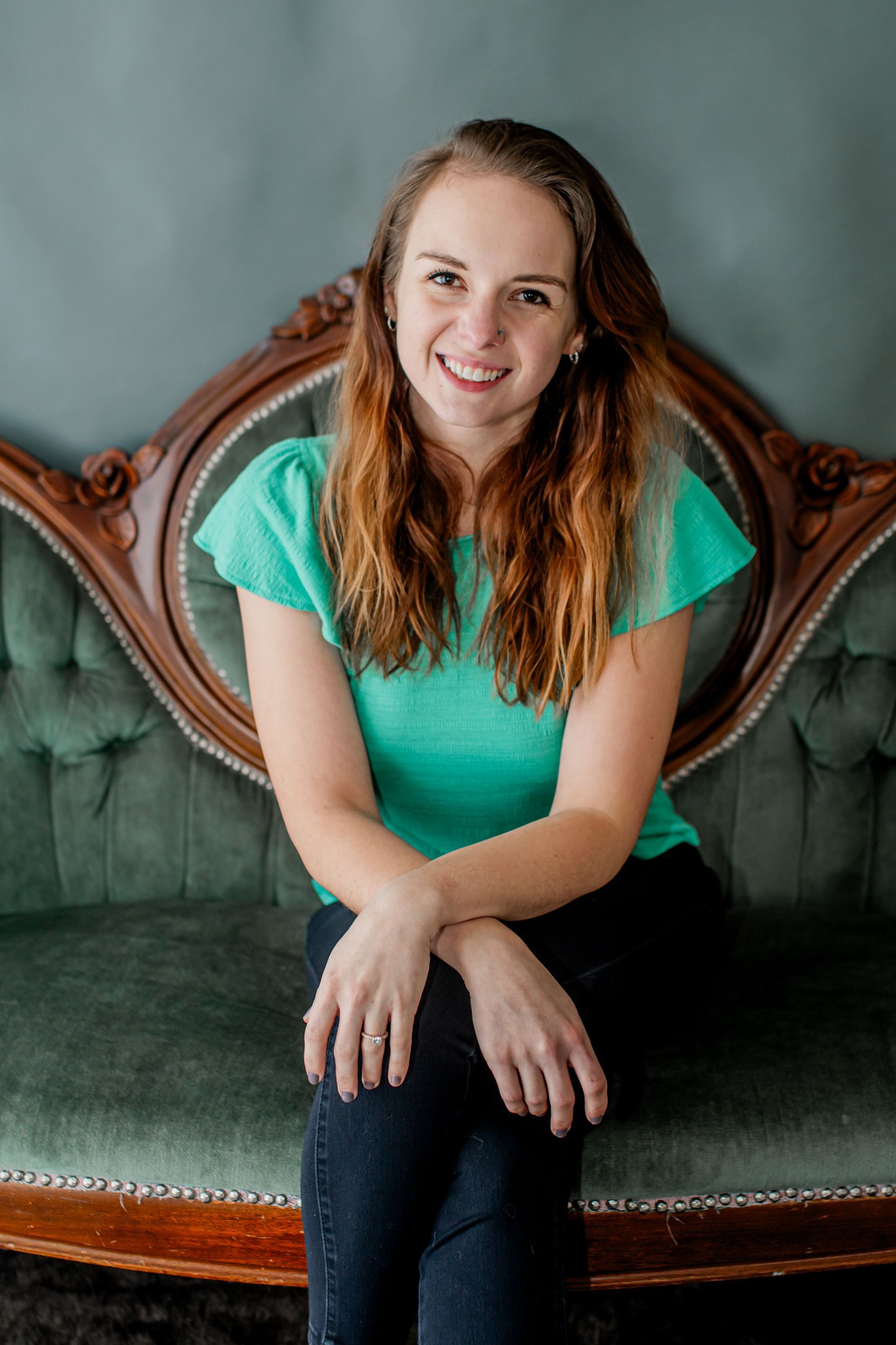 Woman in a teal top sitting on a green vintage sofa, smiling at the camera.