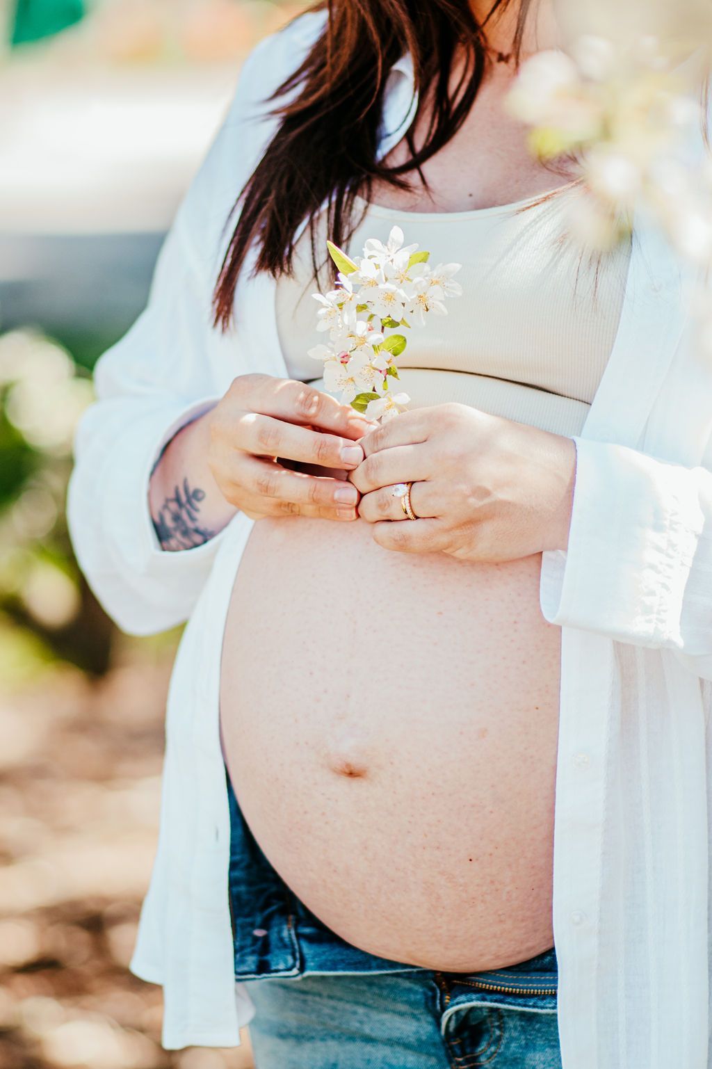 Pregnant person holding small white flowers, wearing an open white shirt and jeans outdoors