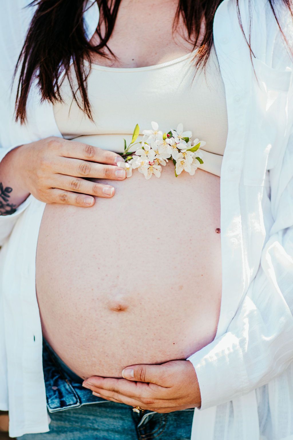 Pregnant person holding white flowers over bare belly in a light sweater and jeans