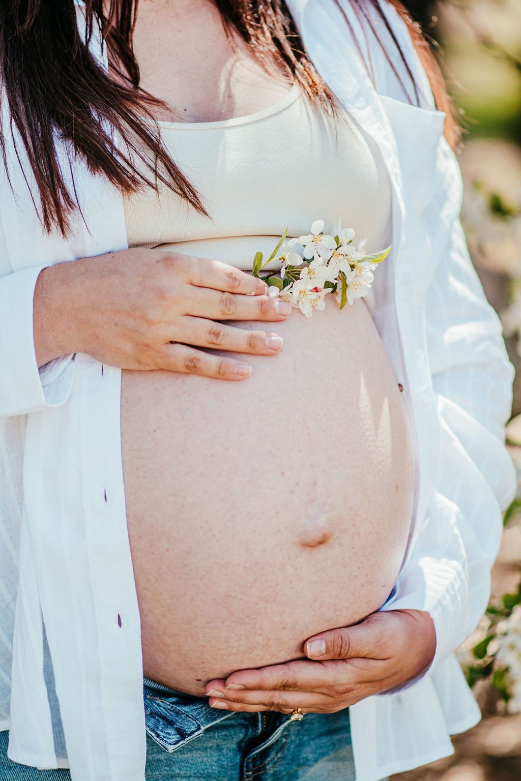Pregnant belly with hands resting on it, holding small white flowers outdoors.