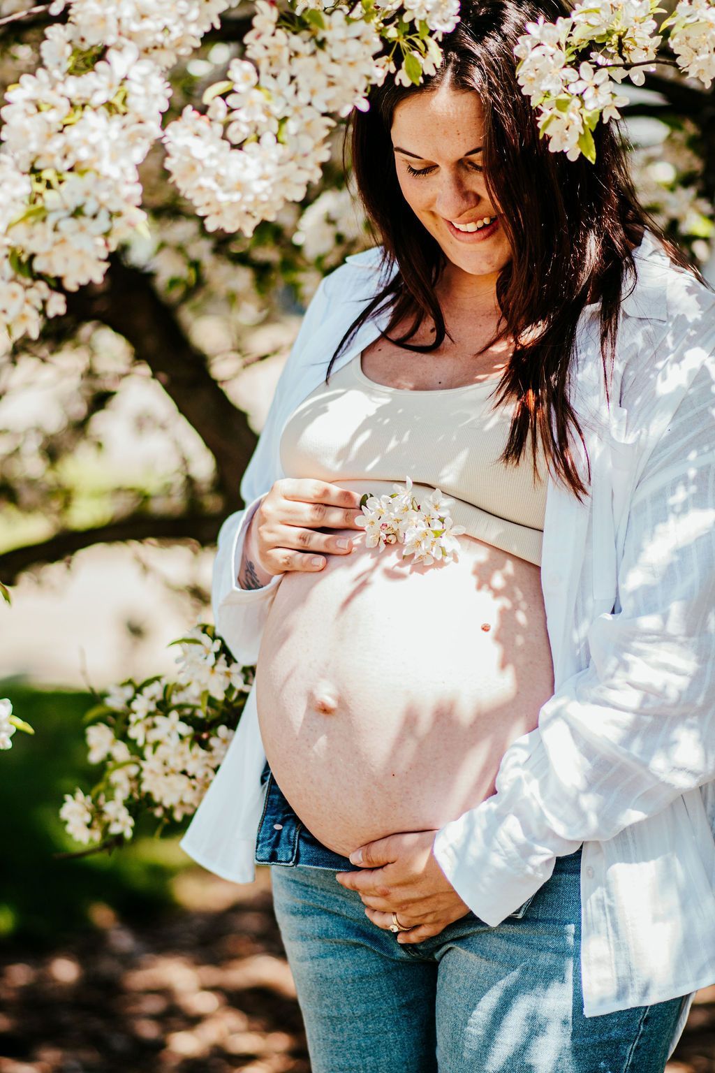 Pregnant person standing under blooming white flowers, smiling and holding their belly outdoors